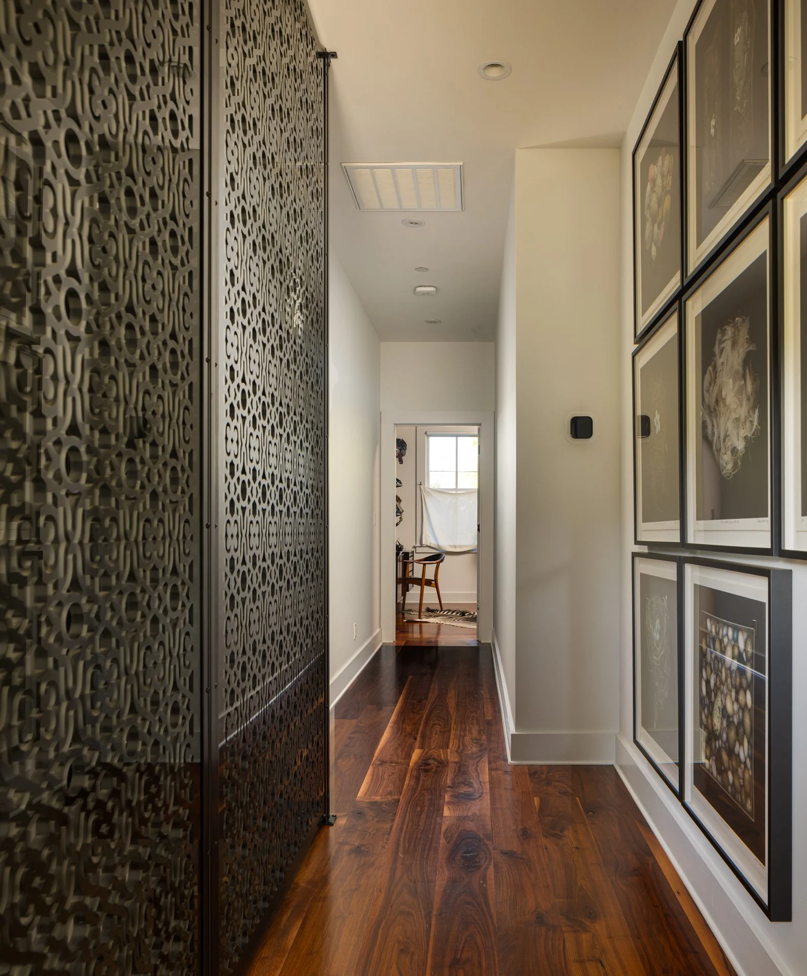 A hallway with dark hardwood flooring, white walls, and a decorative black metal room divider on the left. On the right, there are framed pictures of natural scenes. At the end of the hallway, there is a doorway leading to a room with a chair, a wind