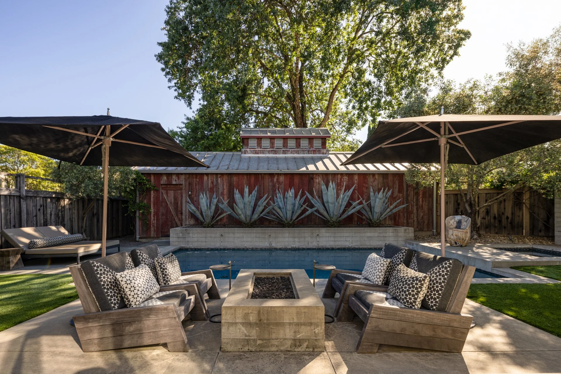 Outdoor patio with a fire feature, surrounded by lounge chairs with pillows, two umbrellas, a swimming pool, a red wooden fence with a row of agave plants, tall trees, and an open sky.