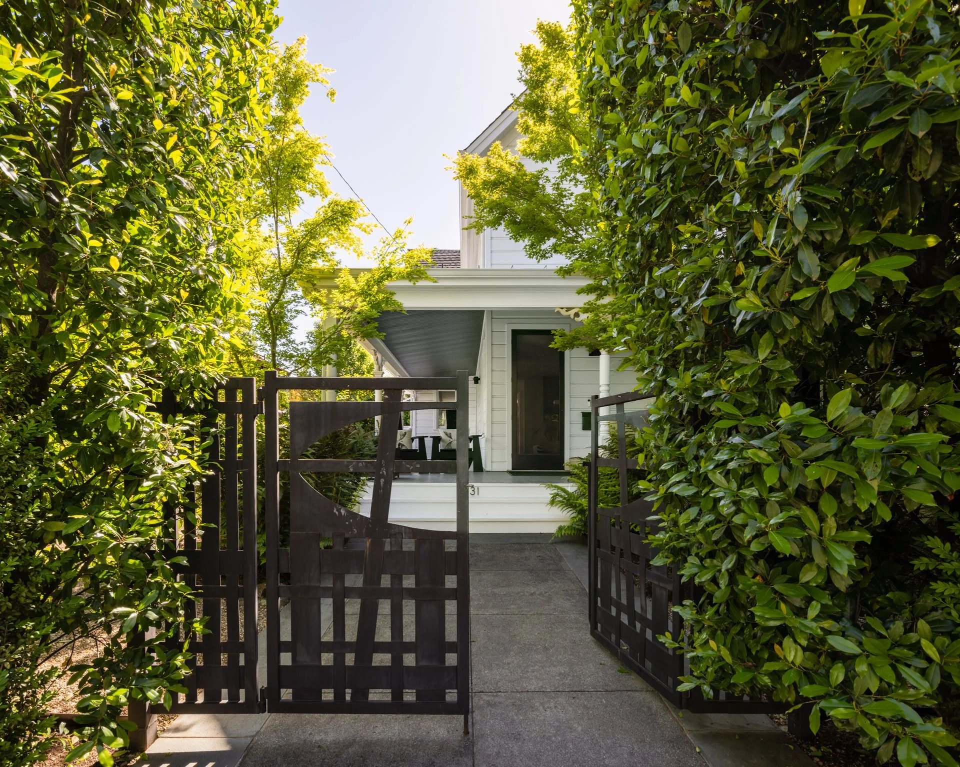 A white house with a porch, partially obscured by green bushes and trees, with a black wooden gate in front and a concrete walkway.