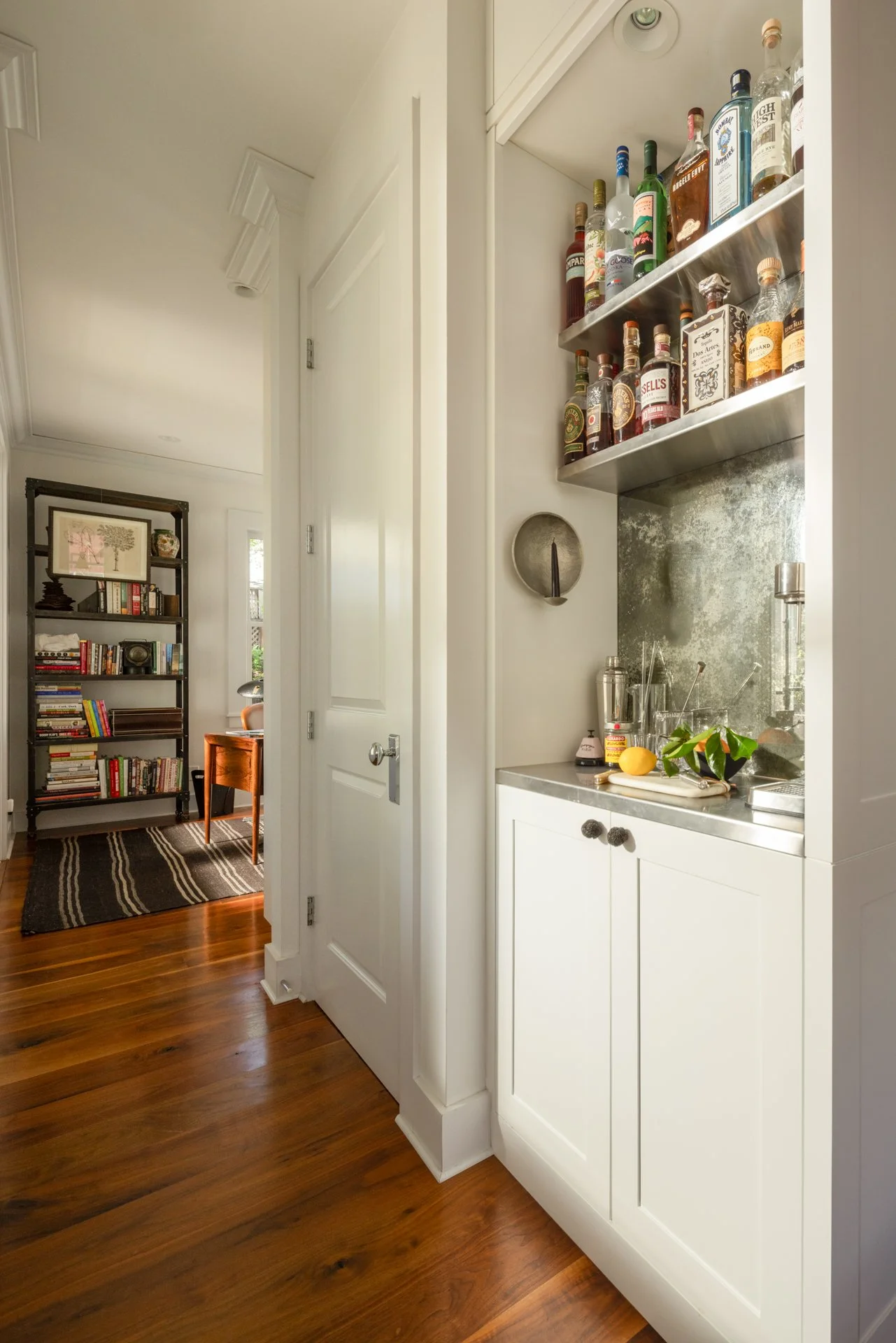 Interior view of a kitchen with white cabinetry, a small countertop with a lemon and green plant, and a shelf above holding various liquor bottles. Adjacent to the kitchen is a room with a bookshelf, a chair, and a patterned rug on wooden flooring.