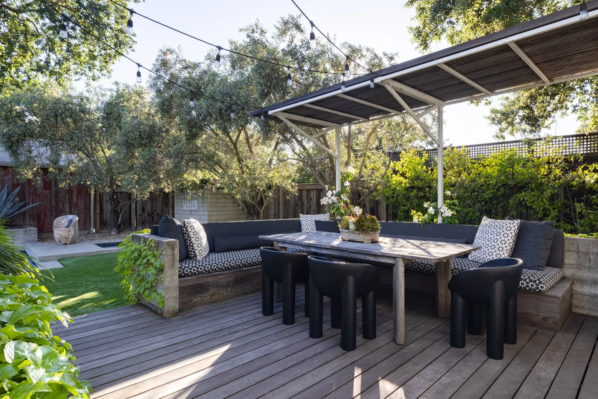 Backyard patio with outdoor seating, a wooden dining table, black chairs, a sectional sofa with patterned throw pillows, string lights overhead, trees, and lush green plants surrounding the space.