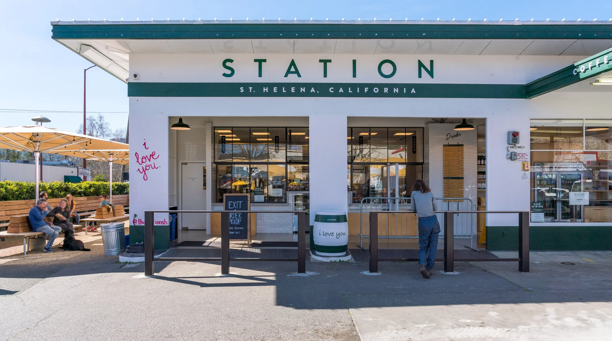 A small train station in St. Helena, California with people sitting on benches and waiting outside. The station has a white exterior with green accents, large glass windows, and a sign that reads 'STATION,' along with the location. There are umbrella