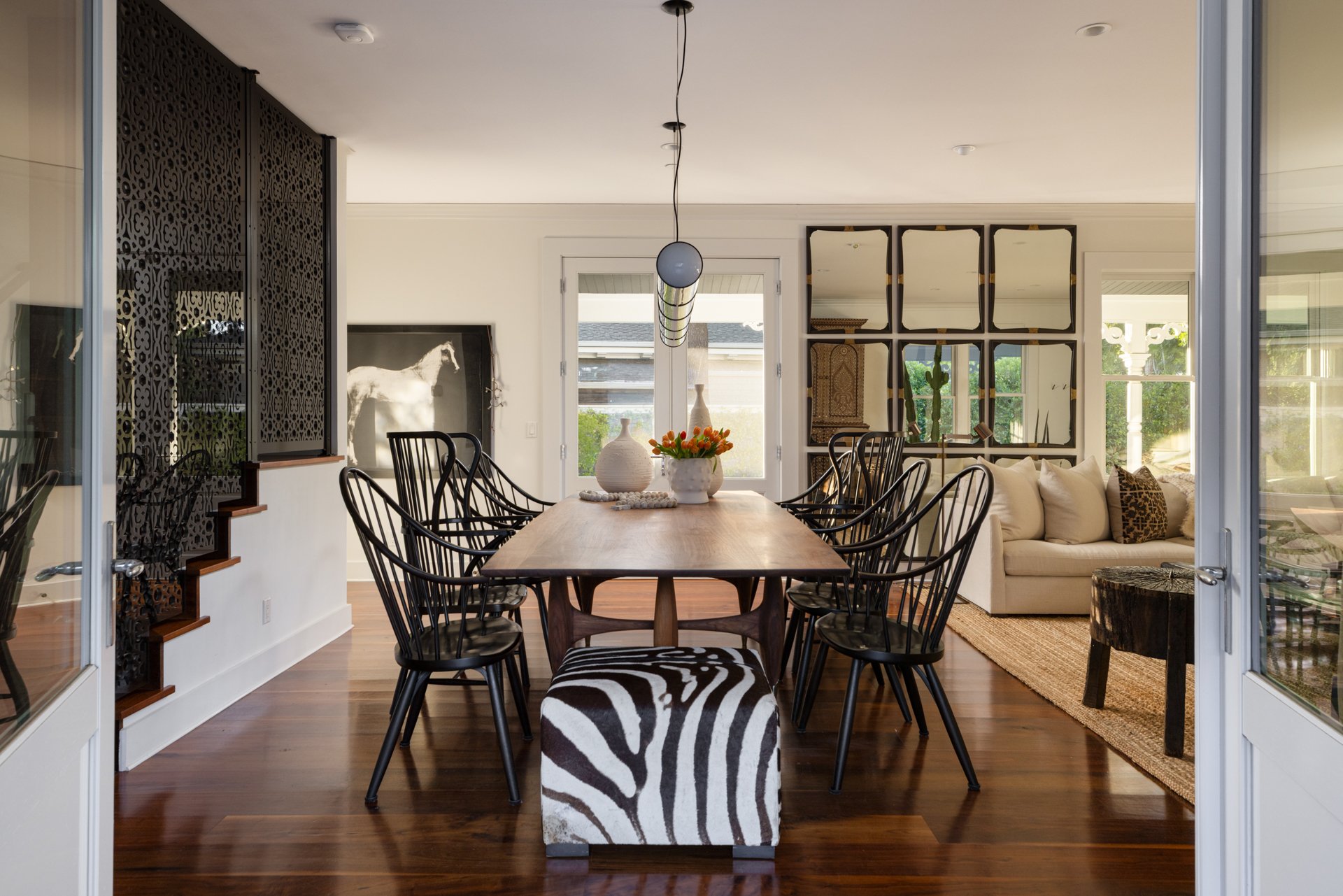 A dining room with a wooden table surrounded by black chairs, zebra print bench, white sofa with pillows, decorative mirrors on the wall, vases with flowers, and large windows showing greenery outside.