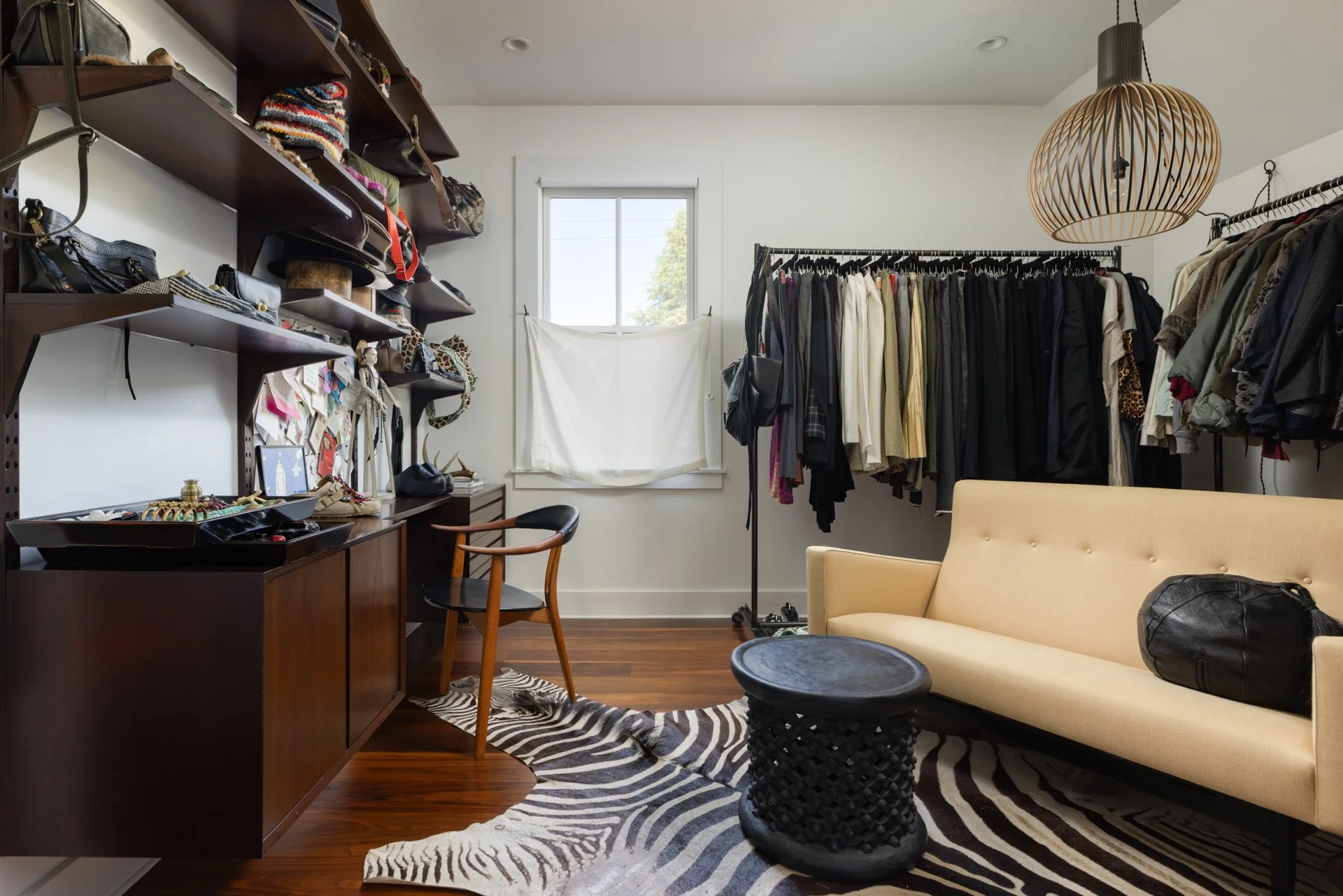 A room with shelves filled with handbags, a desk with jewelry and accessories, a black chair, a hanging rod with clothes, a beige sofa, a black leather bag, a zebra-print rug, and a window with blinds.