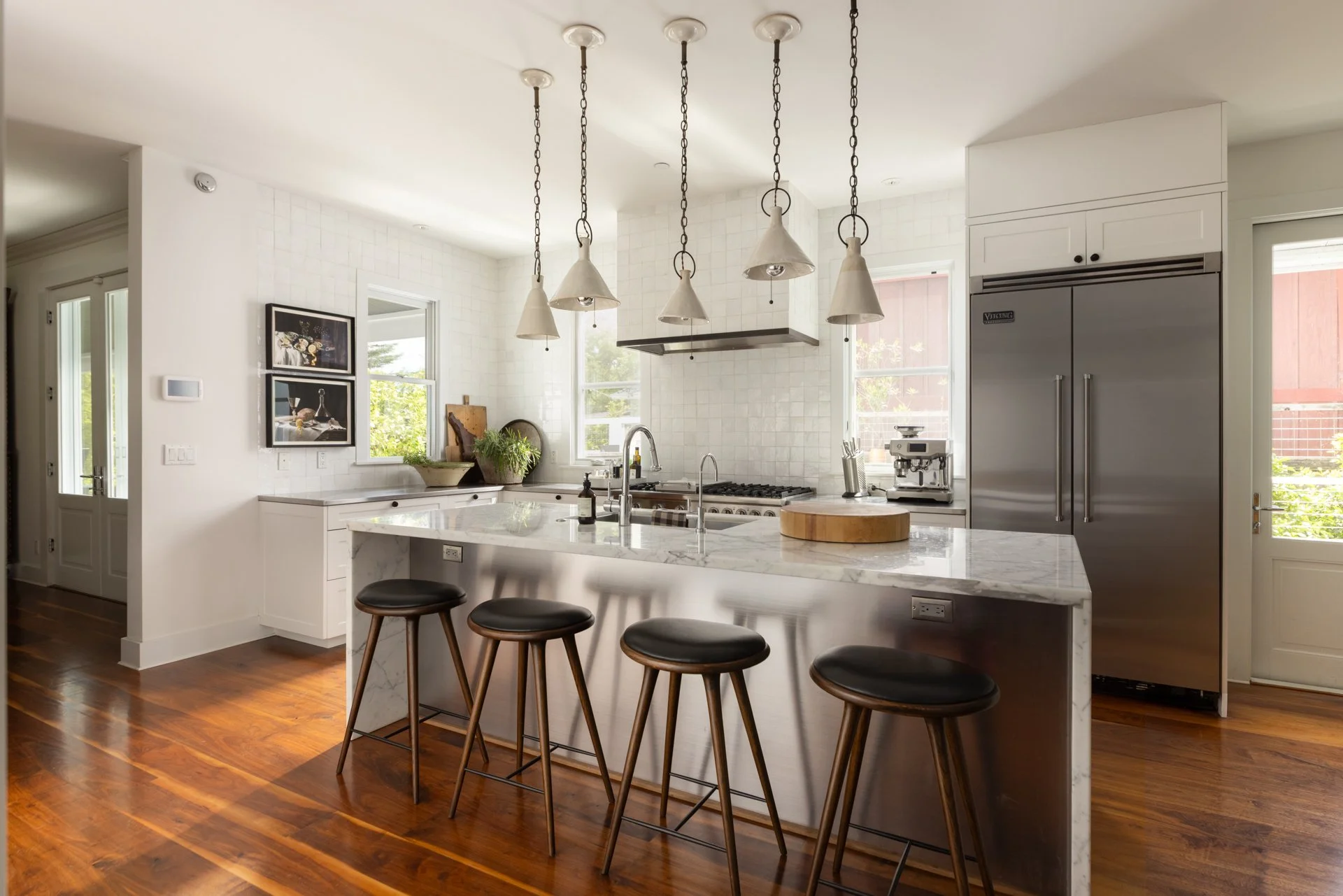 Modern kitchen with white cabinets, marble island, four pendant lights, stainless steel refrigerator, stove, and a small seating area with three stools and a wooden countertop.