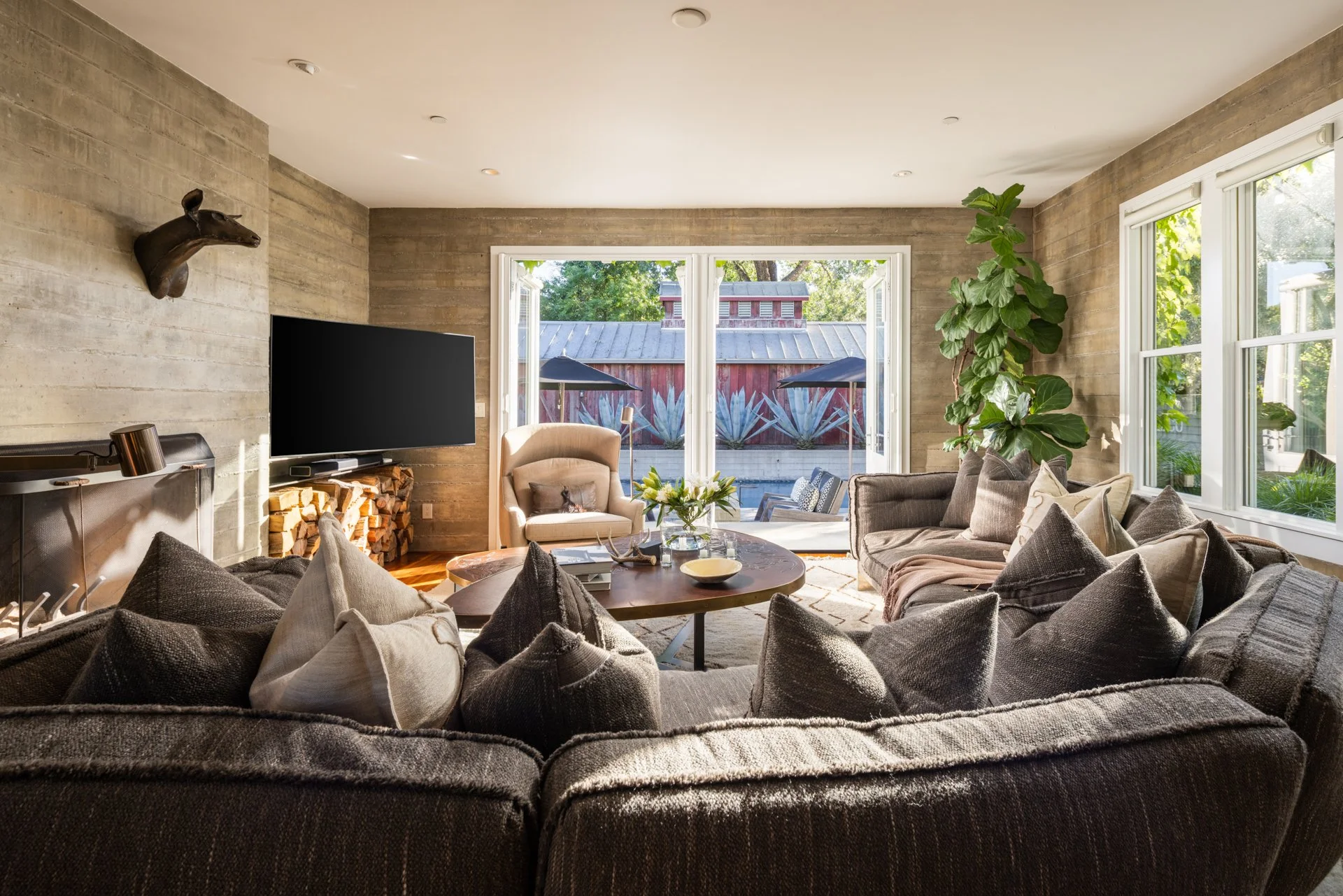 Living room with a large sectional sofa with numerous pillows, a round wooden coffee table, a beige armchair, a flat-screen TV, a wood-burning fireplace, a tall plant near the window, and a view of the backyard with umbrellas and a fence.