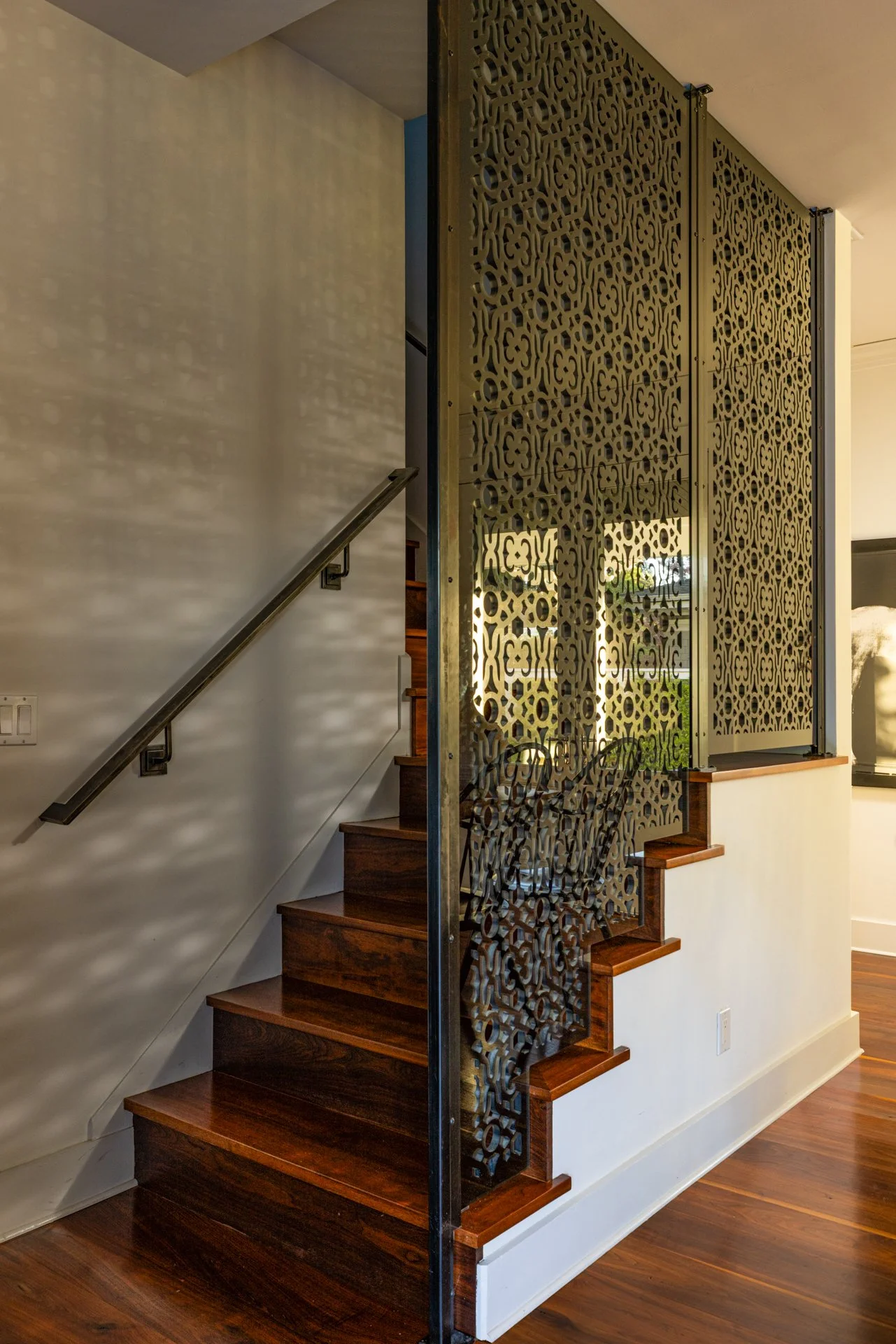 Interior staircase with dark wooden steps, a metallic handrail on the left wall, and a decorative metallic privacy screen with intricate patterns on the right side. The wall on the left is painted white, with light casting shadows from the screen. Th