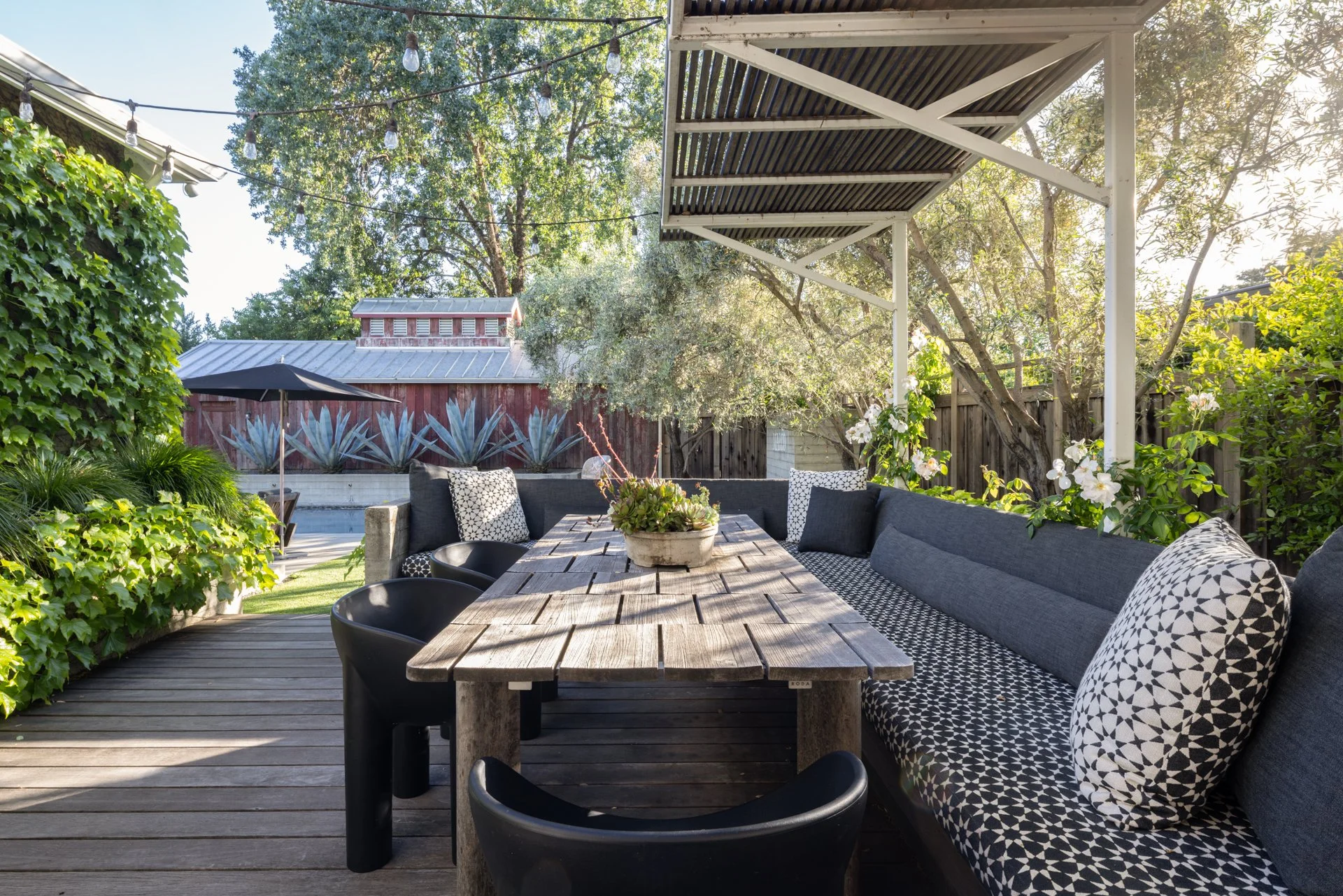 Outdoor patio with a wooden table, black chairs, a gray sectional sofa with patterned pillows, and lush greenery with trees and plants in the background.