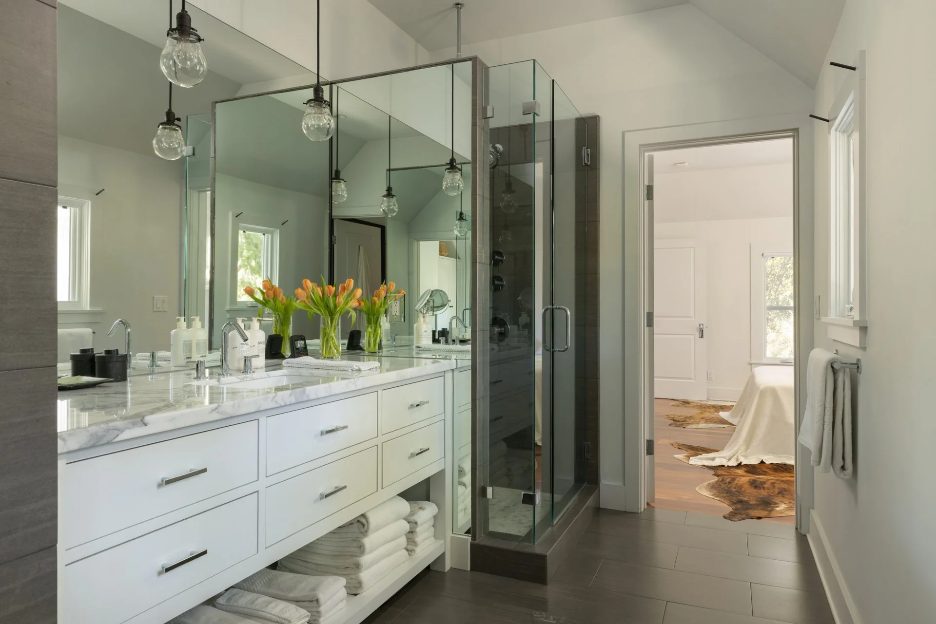 Modern bathroom with white vanity, marble countertop, large mirror, glass shower enclosure, and a visible bedroom through an open door.