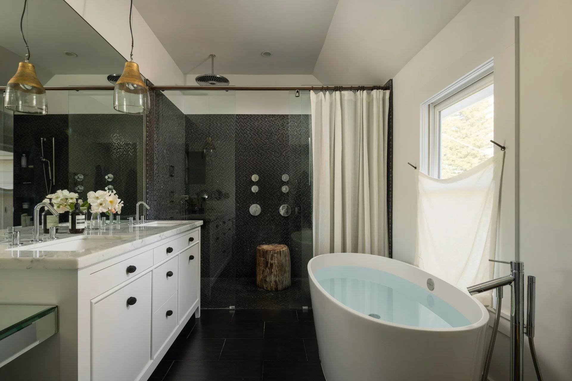 Modern bathroom with a white vanity, black knobs, and a marble countertop, holding a soap pump and flowers. A freestanding bathtub is positioned near a window with a white curtain. The shower area has black mosaic tiles and modern fixtures, with a wood stump as a stool. Two pendant lights with glass and gold accents hang from the ceiling.