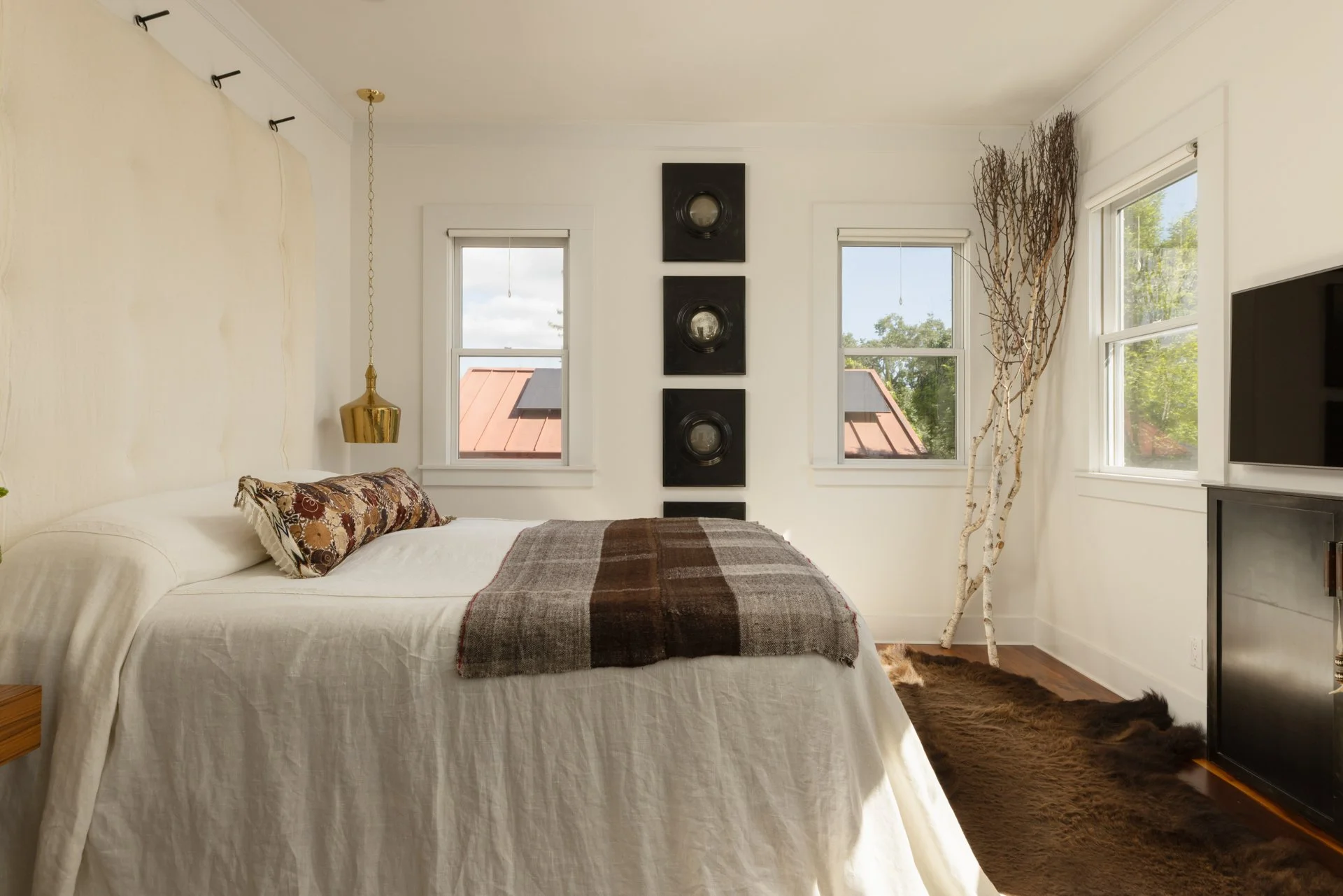 A bedroom with a bed, decorative pillows, a blanket, windows, a wall hanging, and natural light.