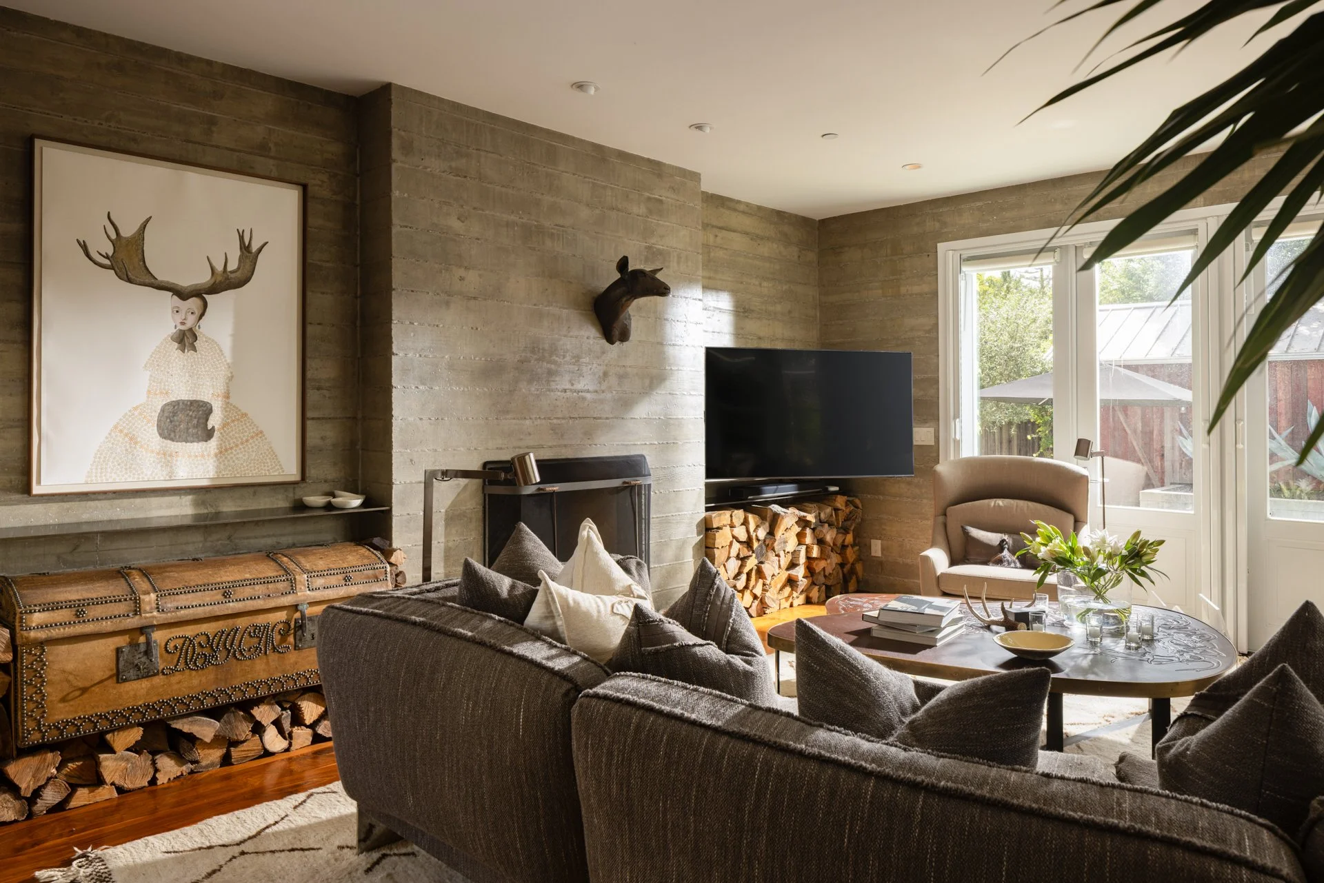 Living room with modern decor, a large TV, a wireframe fireplace, a stack of firewood, a painting of a girl with antlers, and a deer head mounted on a concrete wall near a window.
