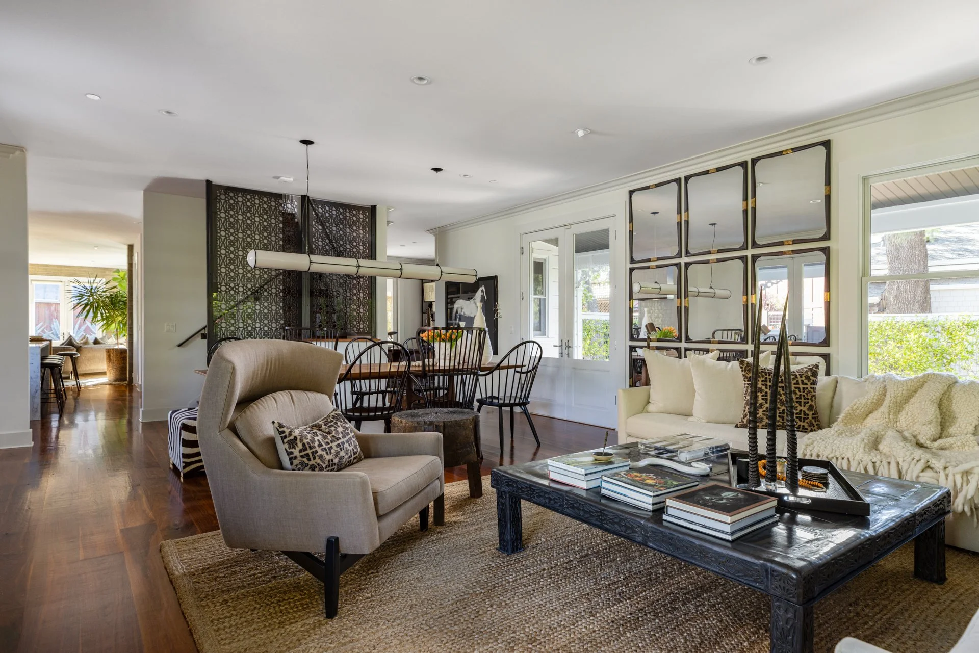 Living room with beige upholstered armchair with patterned pillows, black coffee table with books, a cream sofa with pillows, a large mirror, a lamp, and a view of windows and outdoor greenery.