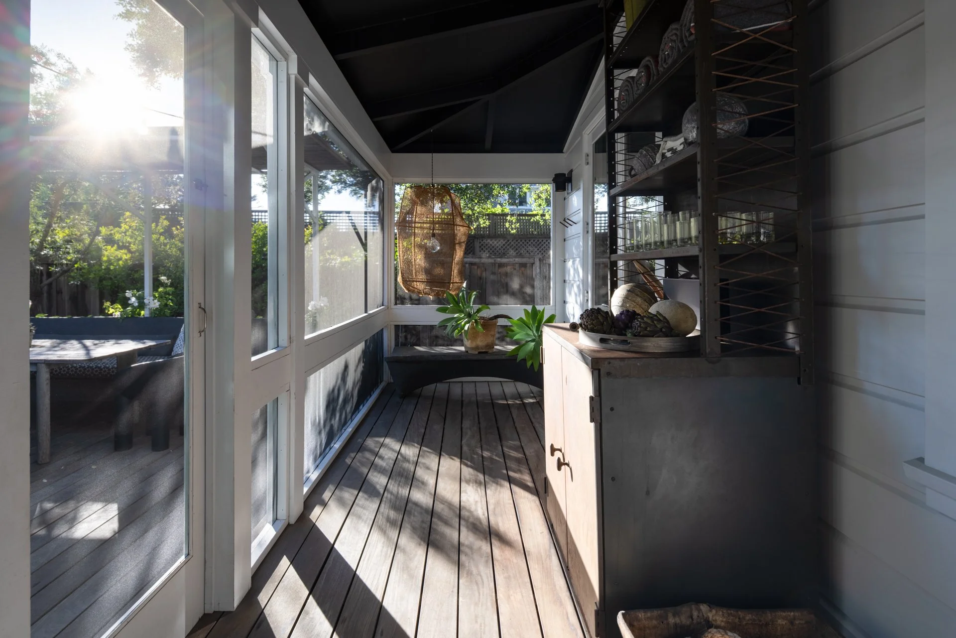 Sunlight shining into a screened porch with wooden flooring, a black metal shelving unit with glassware, a wooden cabinet with artichokes and vegetables, a potted plant, and an outdoor view with trees and a wooden fence.