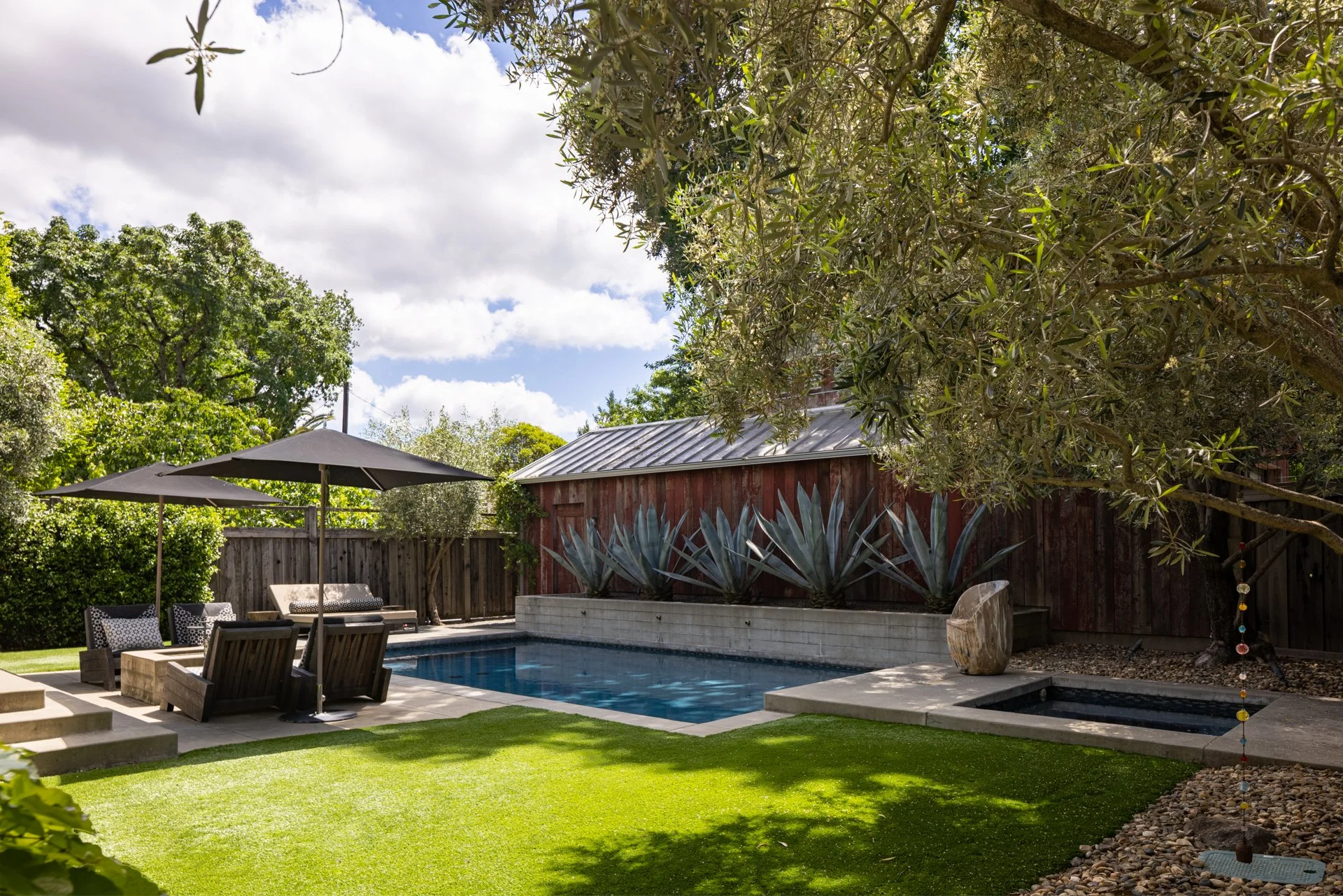 A backyard with a swimming pool, lounge chairs, patio umbrellas, lush green grass, various plants, and trees under a partly cloudy sky.