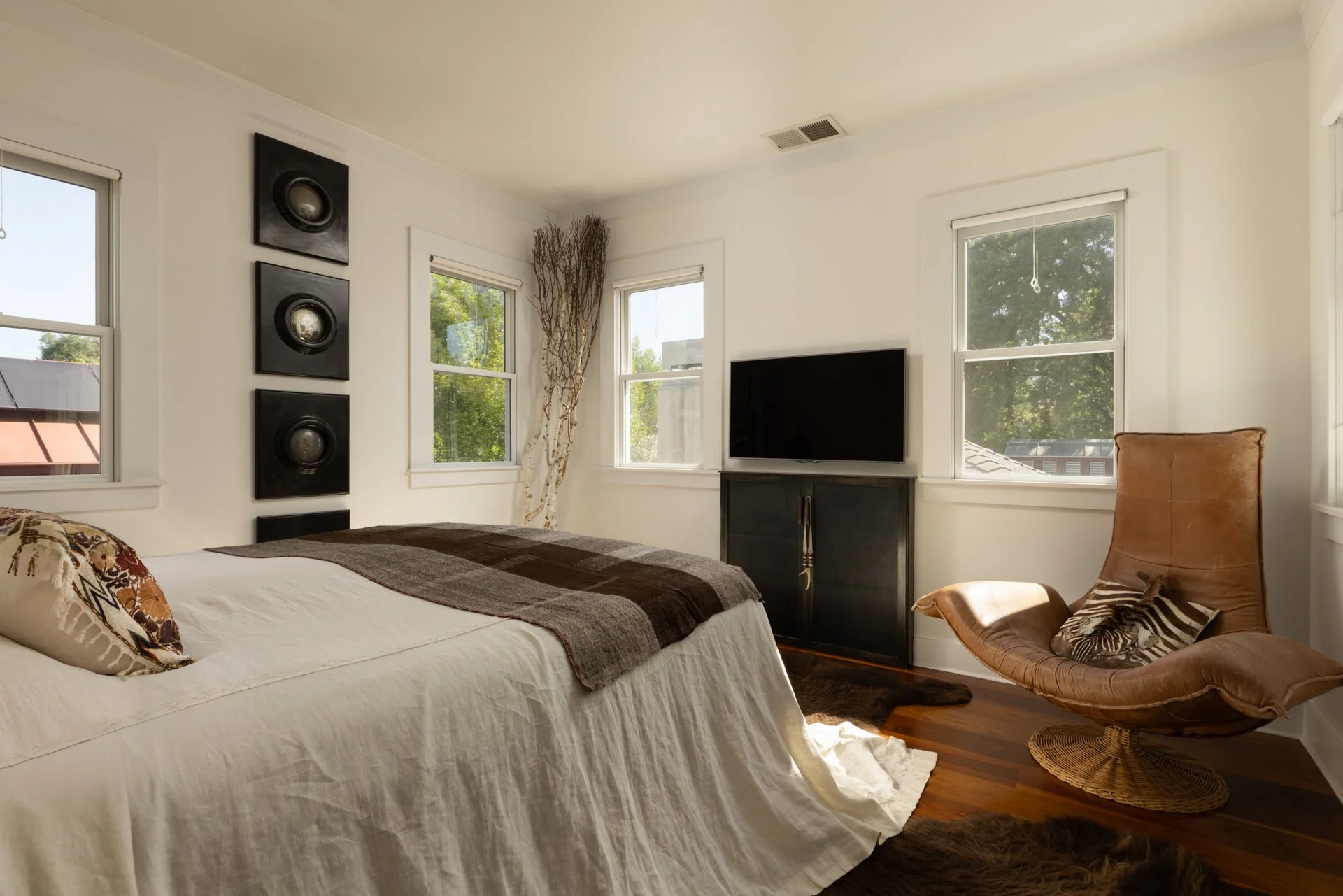 A bedroom with a bed covered in a white sheet and decorative pillows, a black TV on a dark cabinet, a leather chair, and three windows with green trees outside.