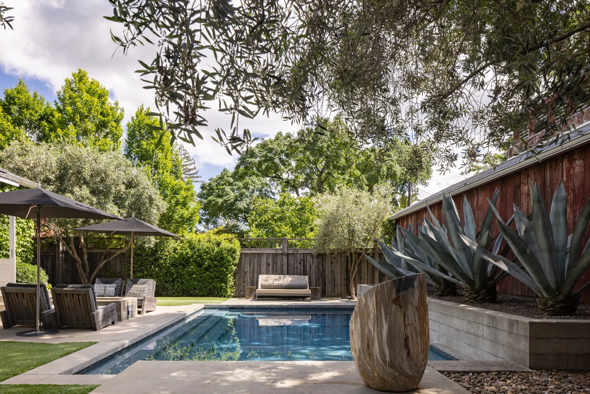 Backyard with swimming pool, outdoor furniture, and large potted plant, surrounded by trees and a wooden fence under a partly cloudy sky.