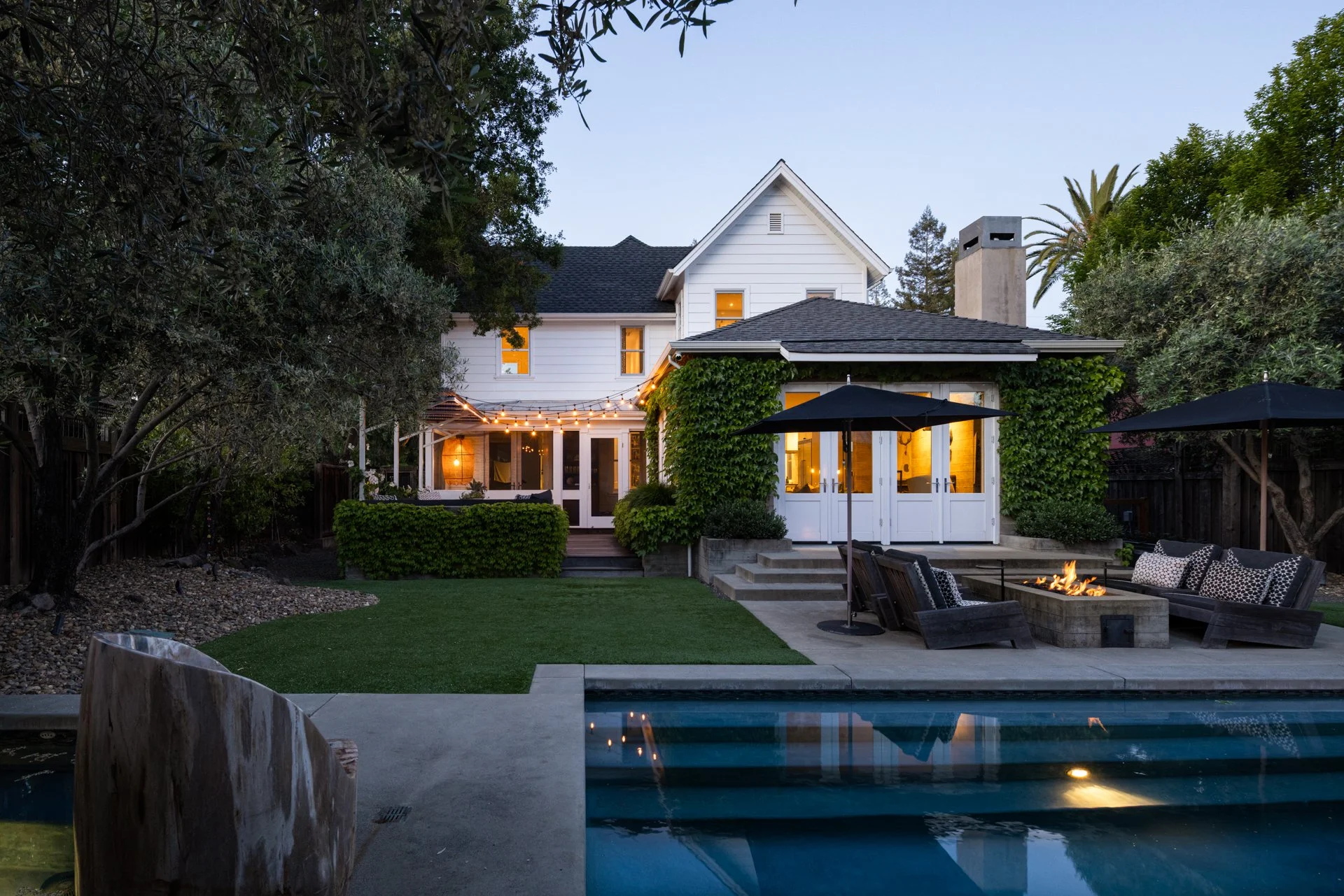 Backyard patio of a house at dusk with string lights, outdoor seating around a fire pit, a swimming pool, and lush green trees.