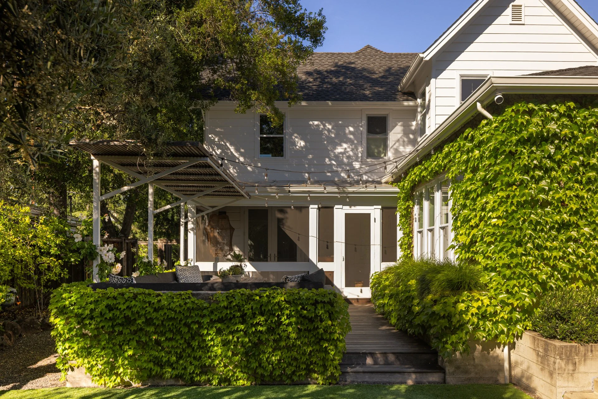 A backyard with lush greenery, including a large ivy-covered wall, a wooden deck, a patio area with outdoor furniture, and a white house with multiple windows and a sloped roof, under a sunny blue sky.