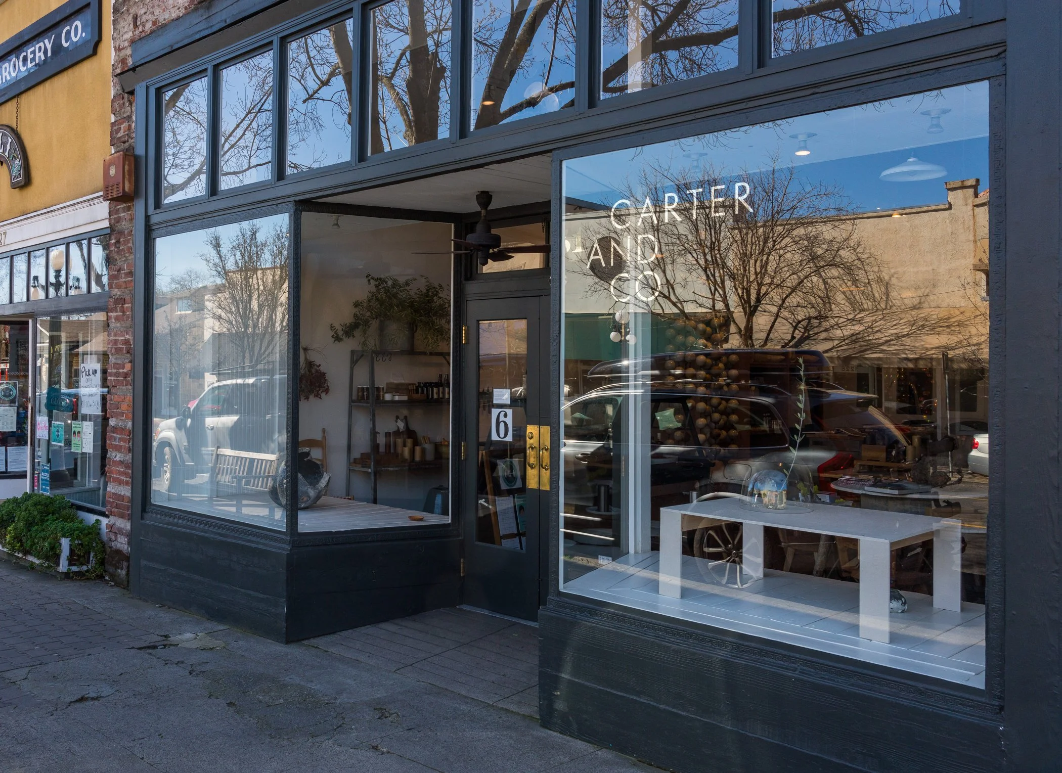Exterior of a store called Carter and Co with large glass windows reflecting trees and the sky. The store has a black facade with a brass-colored door handle and various decorations inside. The street outside has parked cars and a storefront with a s