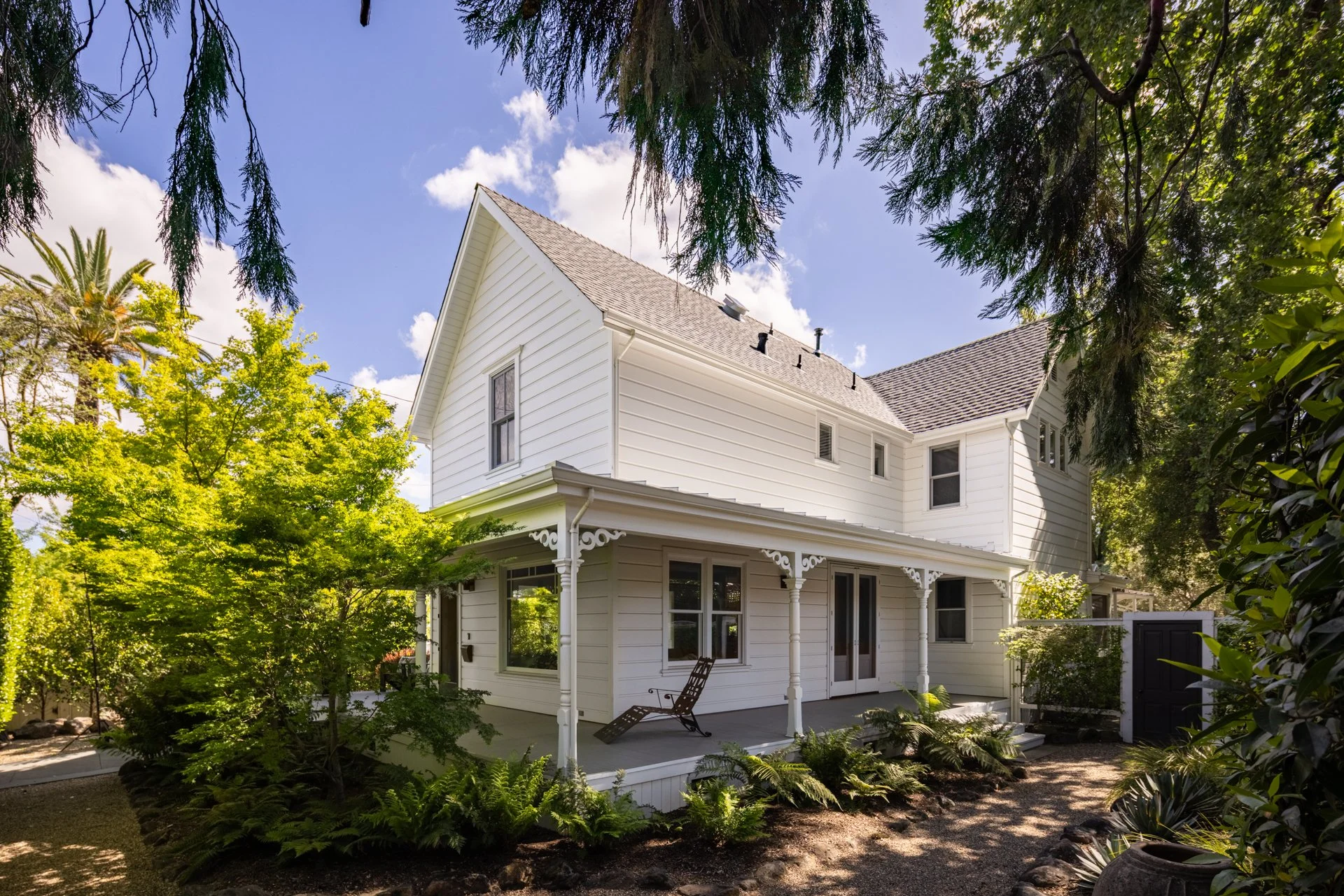 White two-story house with porch, surrounded by green trees and plants, under a partly cloudy sky.