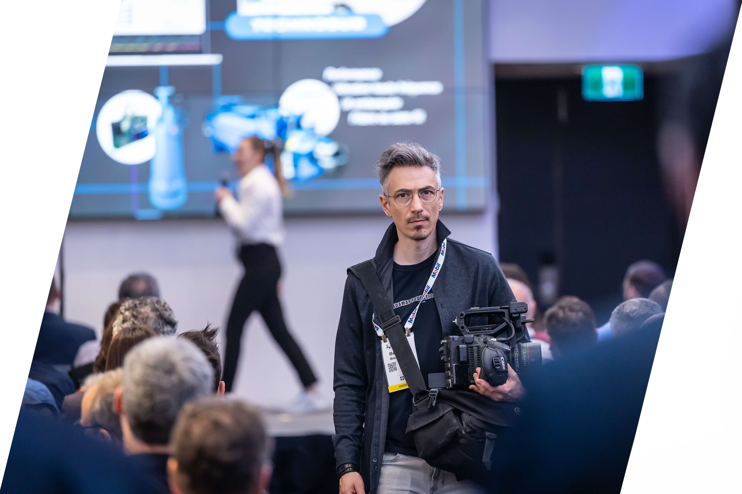 Bernard Boulanger portant un appareil vidéo professionnel dans une salle pleine de personnes, devant un grand écran digital.