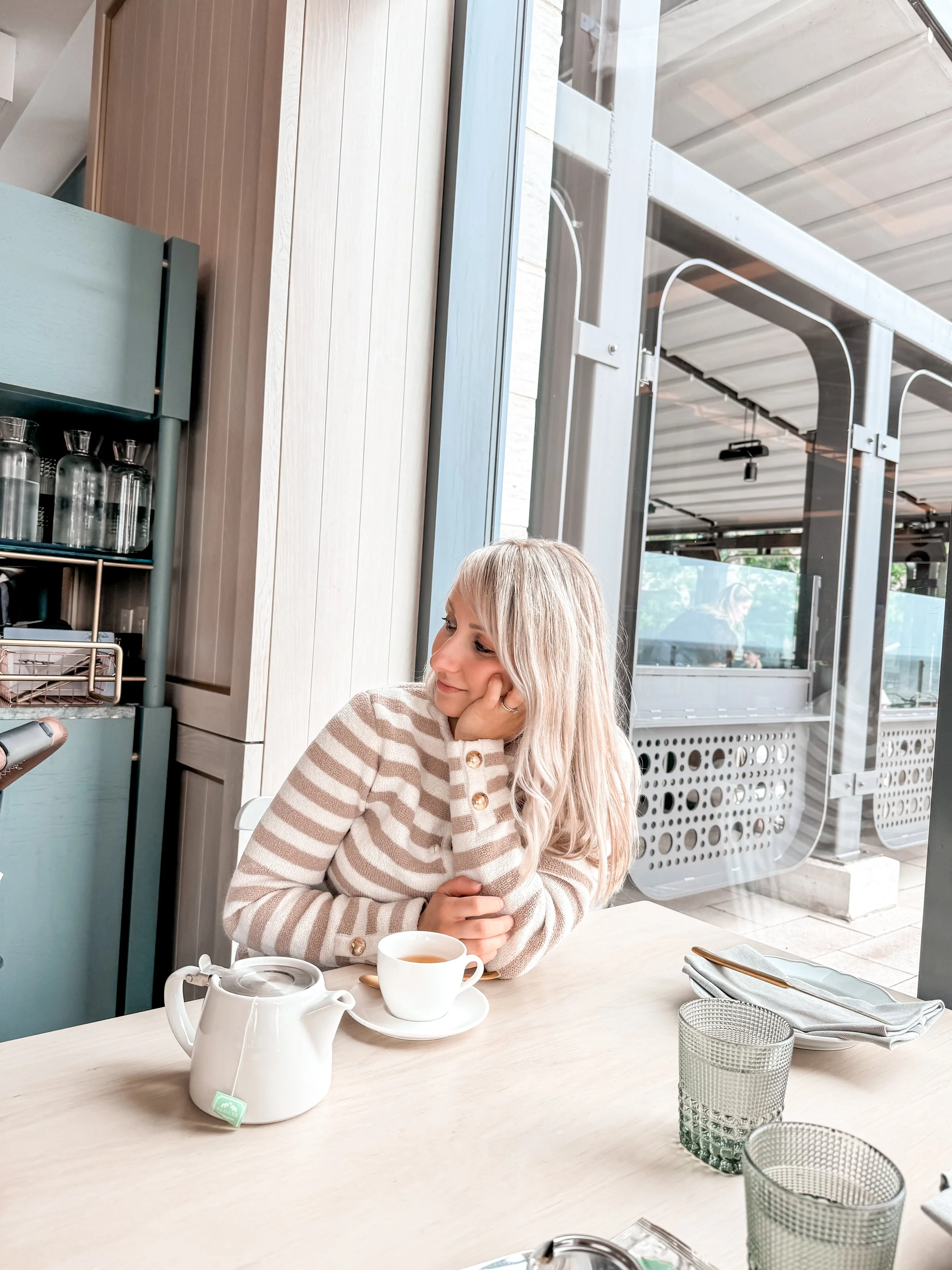 A woman sitting at a light-colored table in a cafe, wearing a beige and white striped sweater, with her head resting on her hand and smiling slightly. On the table, there is a white teapot, a cup of tea, and some glass cups, with a set of silverware wrapped in a napkin nearby. The cafe has large windows, revealing a reflection of people outside.