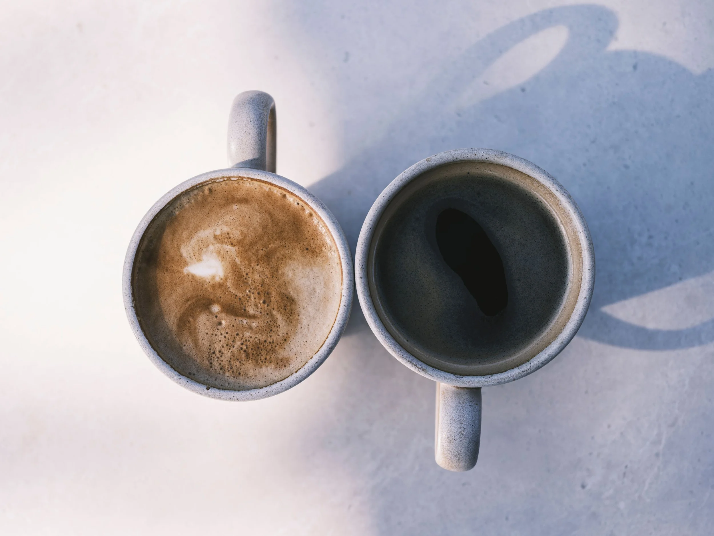 Two ceramic mugs filled with coffee, one with latte art, on a light-colored surface, with shadows cast by the mugs.