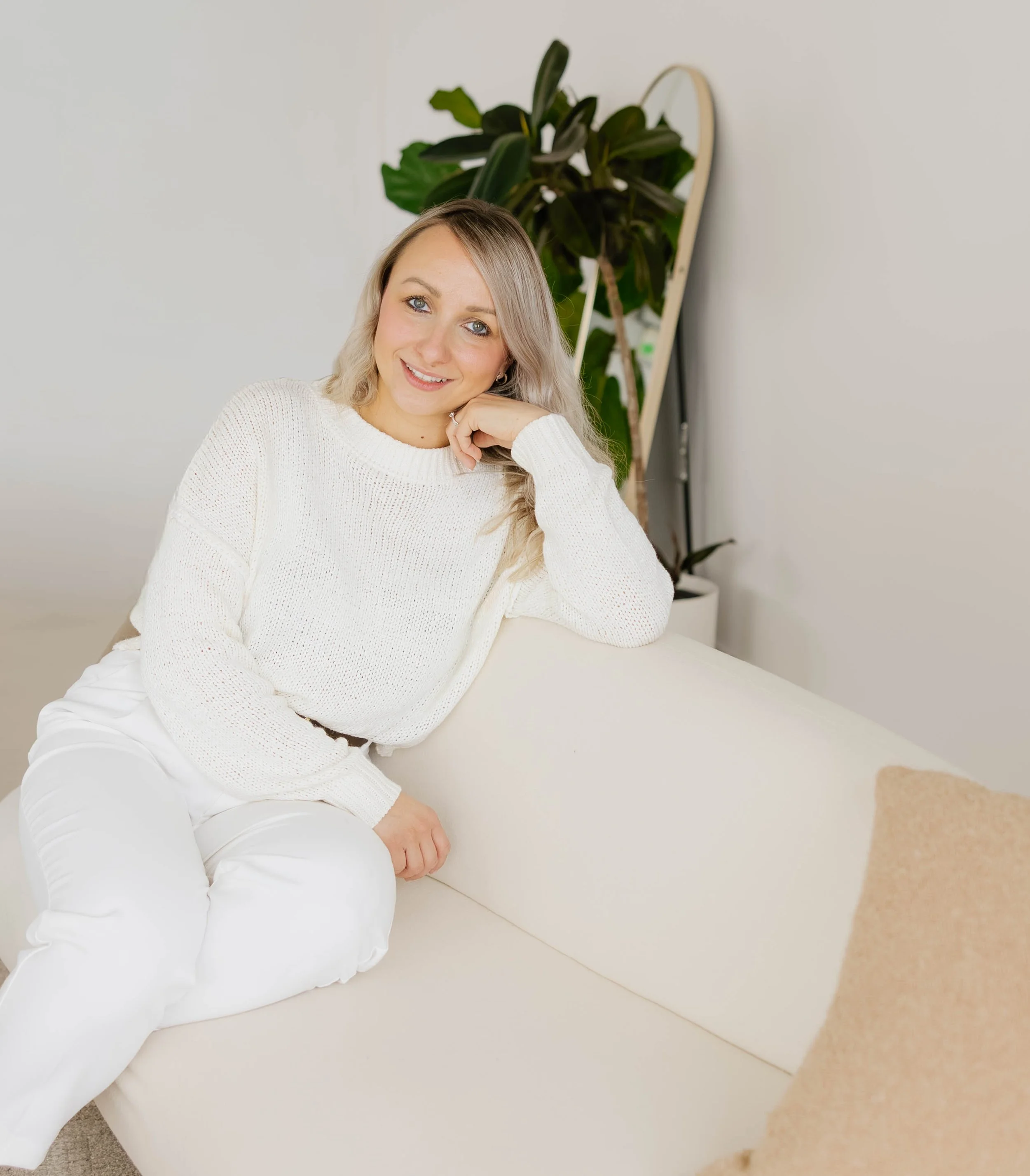 A young woman with blonde hair and blue eyes wearing a white sweater and white pants sitting on a beige couch inside a room with a white wall and a tall green plant in the background.