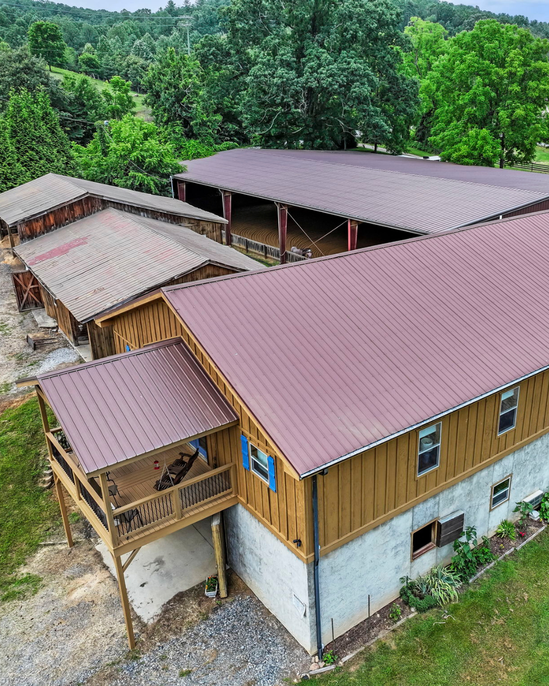 Aerial view of barn house with a red metal roof, wooden siding, and a balcony with outdoor furniture. Surrounding trees and farm buildings are visible. 442 Blantyre Church Rd, Horse Shoe, NC