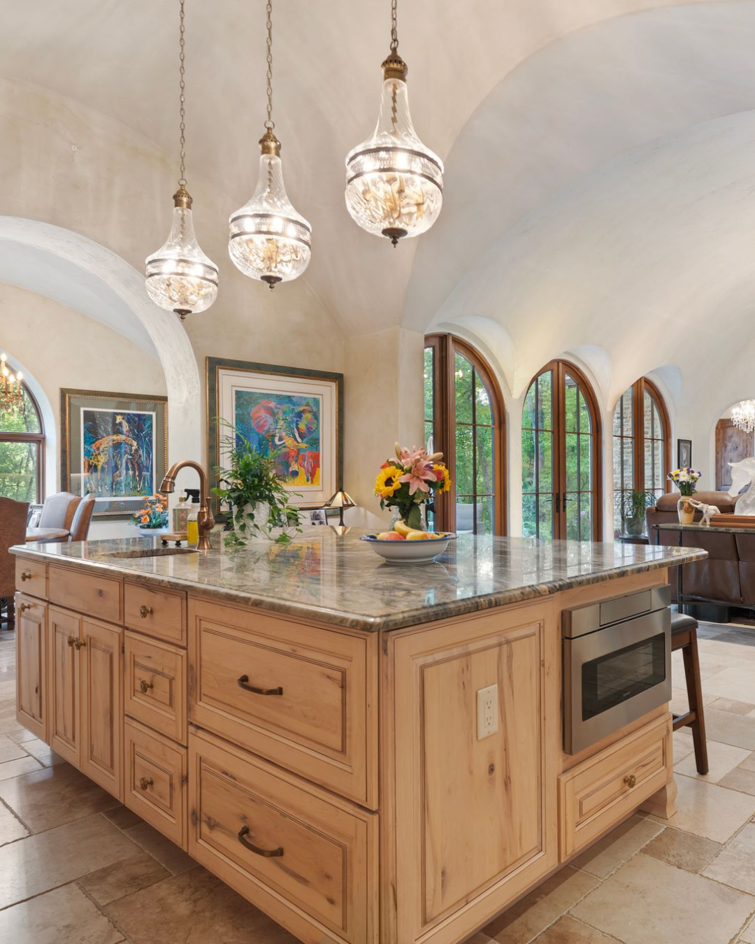 A luxury kitchen with a large island, a marble countertop, and three ornate glass pendant lights hanging above. There are floral arrangements on the island, and large arched windows allow natural light into the space. 959 & 957 Dave Whitaker Rd