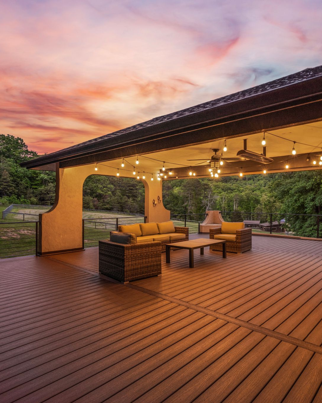 A spacious outdoor balcony with wooden flooring, furnished with wicker sofas, cushions, and a coffee table. String lights are hanging from the ceiling, and the setting overlooks lush green horse pastures at 959 & 957 Dave Whitaker Rd, Horse Shoe, NC