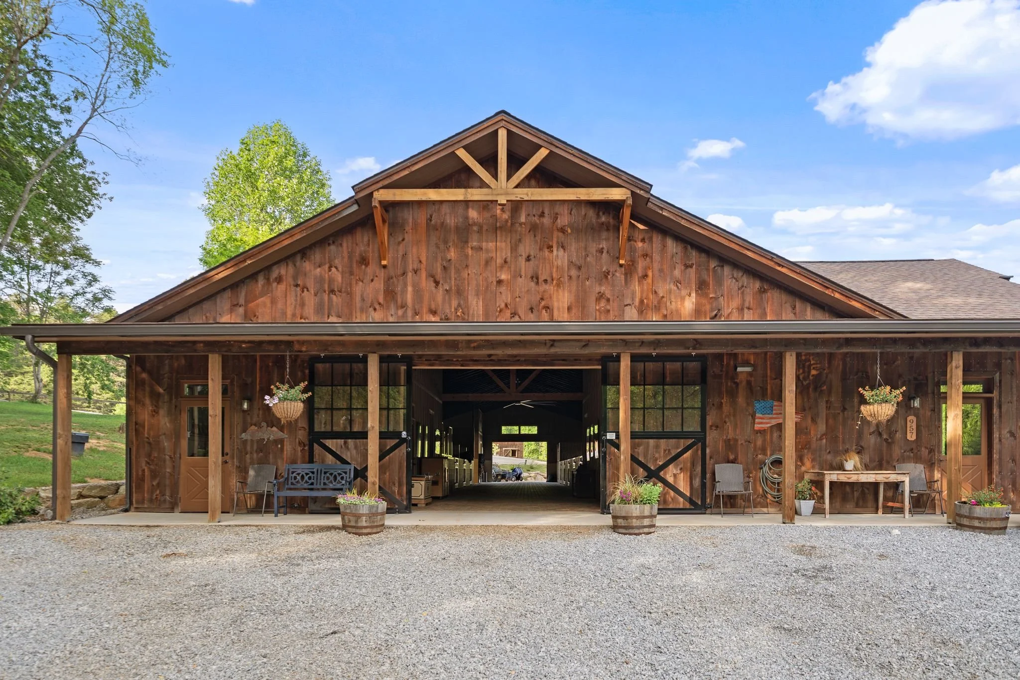 A large wooden barn with an open central area, decorated with hanging flower baskets and outdoor chairs, set against a backdrop of green trees and a blue sky with some clouds.