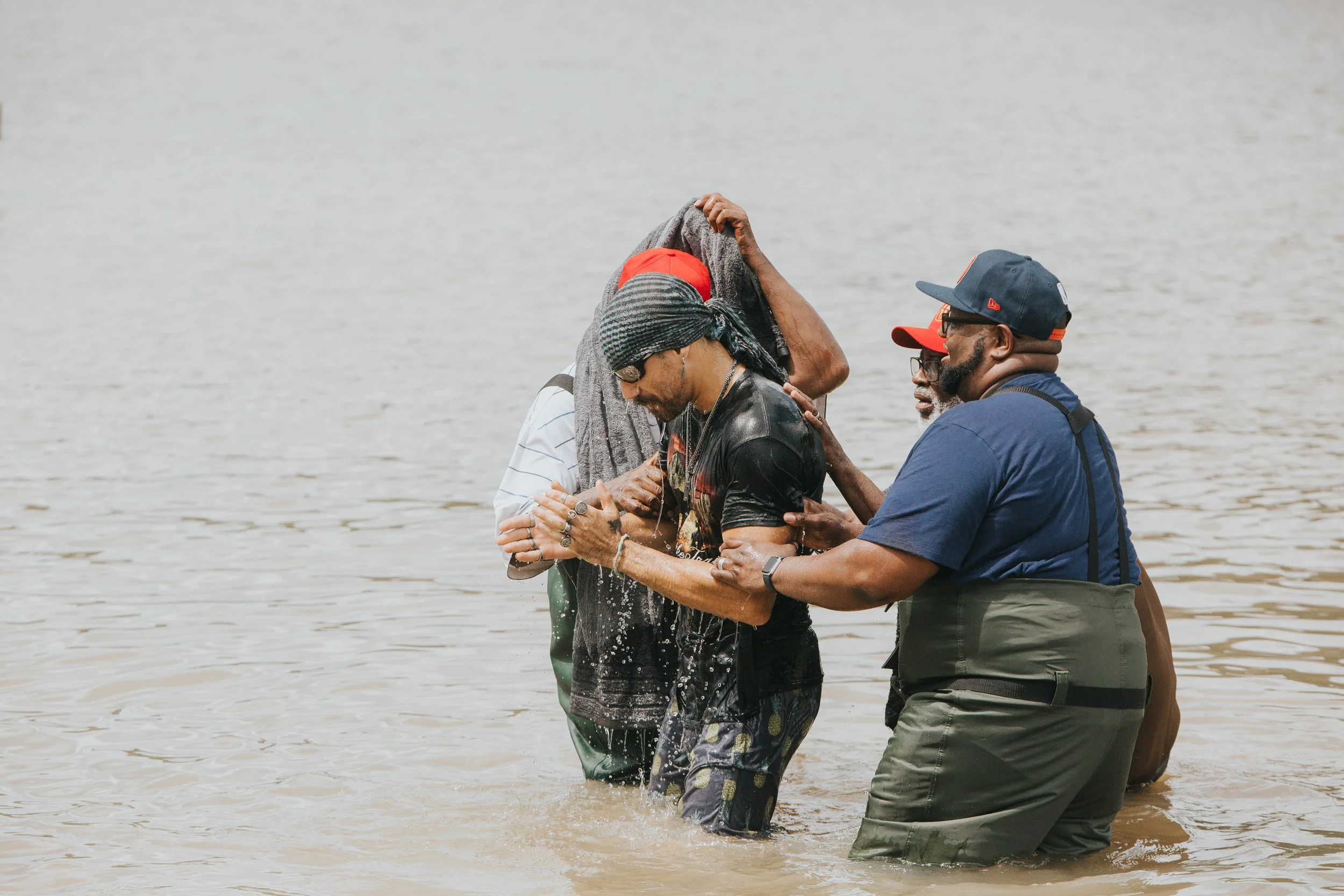 A group of four people standing in water, participating in a baptism ceremony by immersing an individual while others support and assist, with a large body of water in the background.