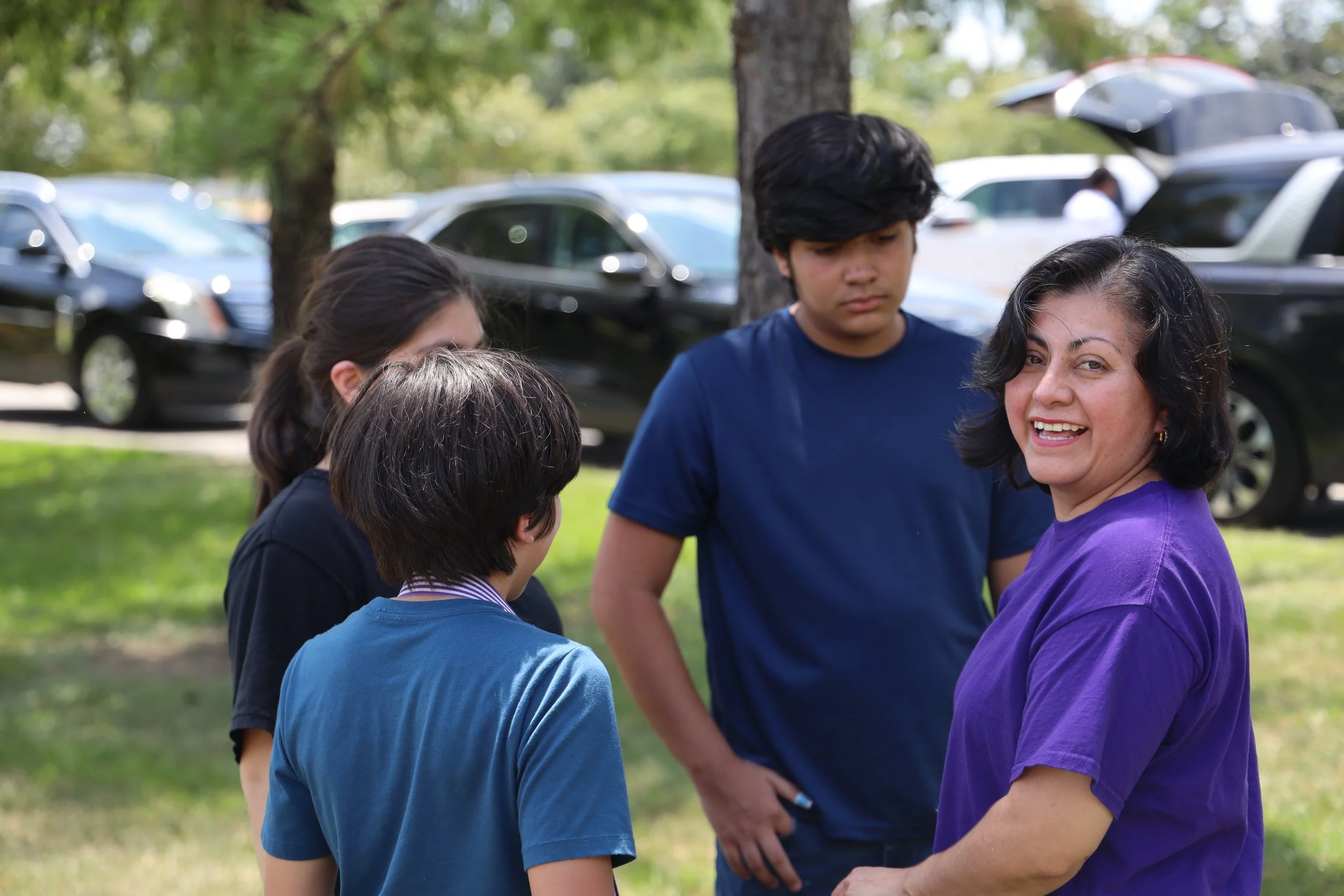 A group of people gathered outdoors under a tree, engaged in conversation. The background shows parked cars and a sunny day.