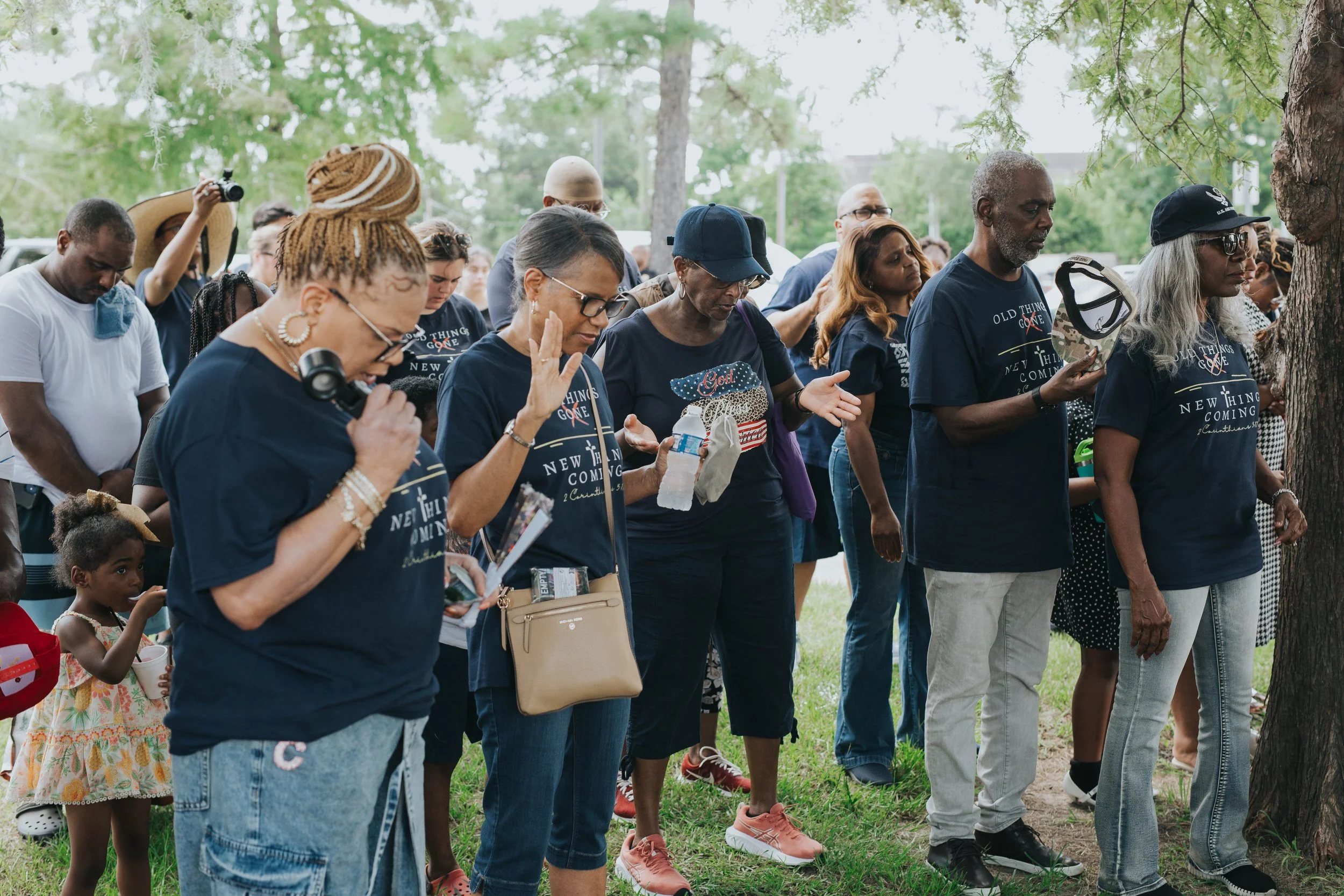 A diverse group of people engaging in prayer or reflection outdoors under trees, some with eyes closed and hands raised, during a community event.
