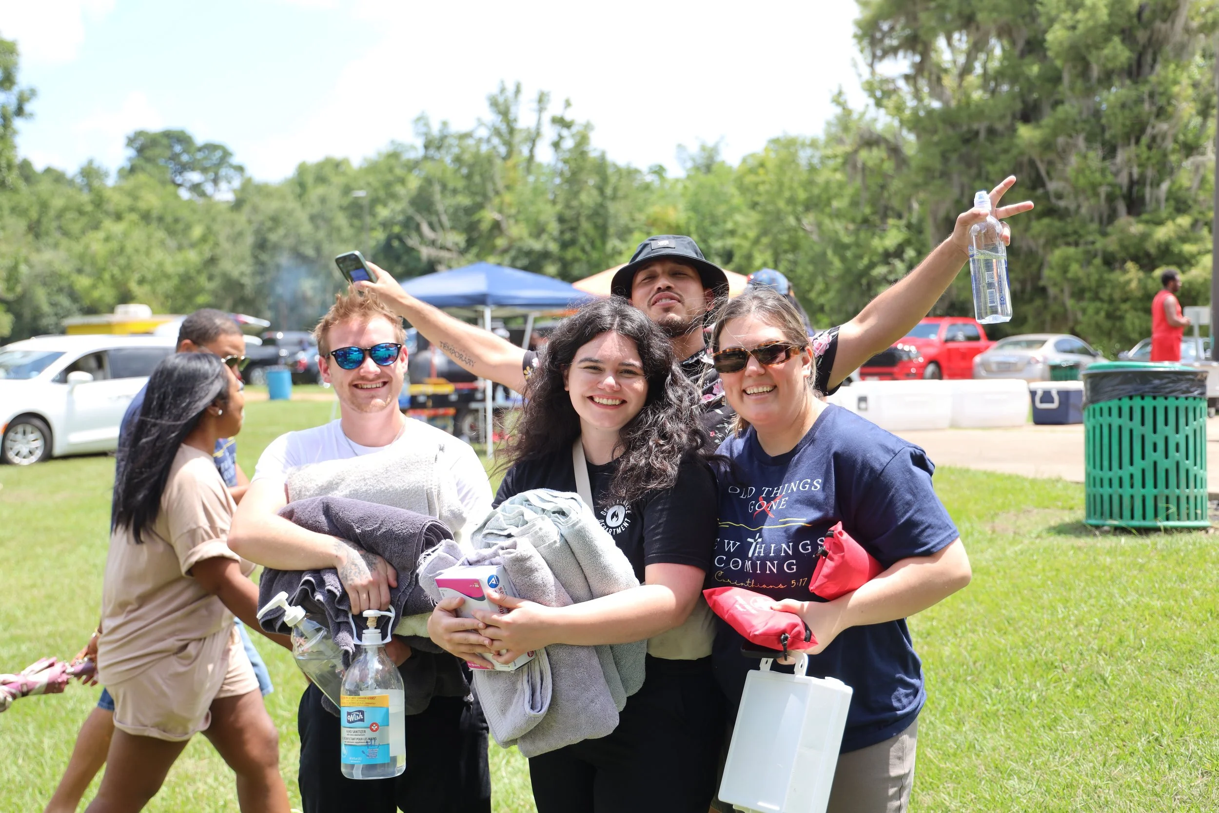 Group of five young people gathered outside on a bright, sunny day, smiling and posing for the camera. There are parked cars and trees in the background, with some tents and people in the distance.