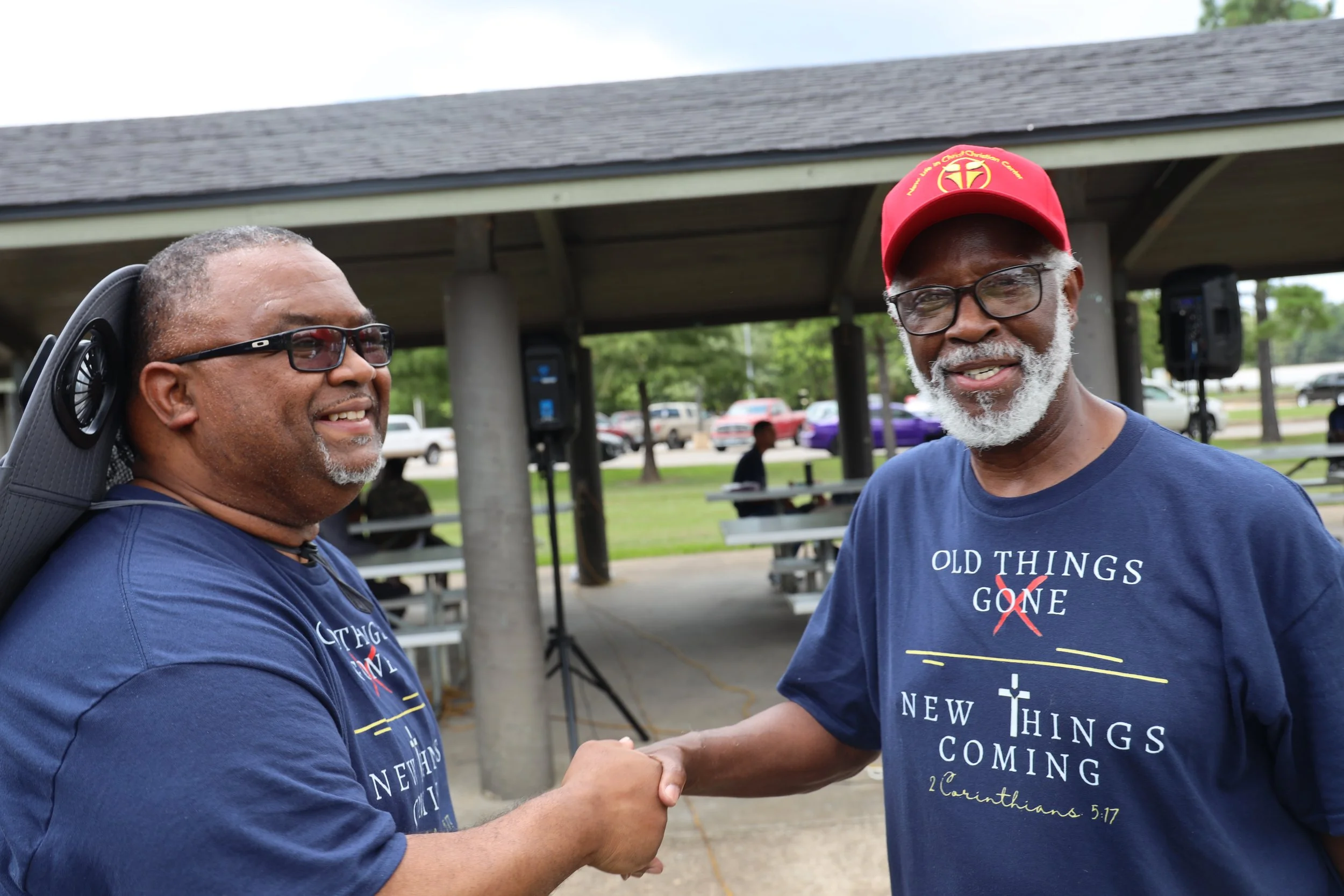 Two smiling men shaking hands outdoors during a community event, both wearing navy blue t-shirts with Christian messages and text, one wearing glasses and a red cap, the other with black glasses and a backpack, with a pavilion and parked cars in the background.