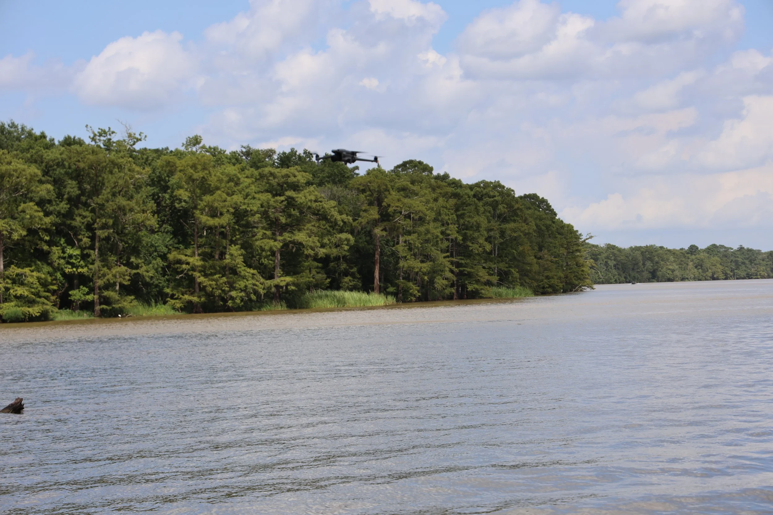 A drone flying over a river with a forested shoreline and a partly cloudy sky in the background.