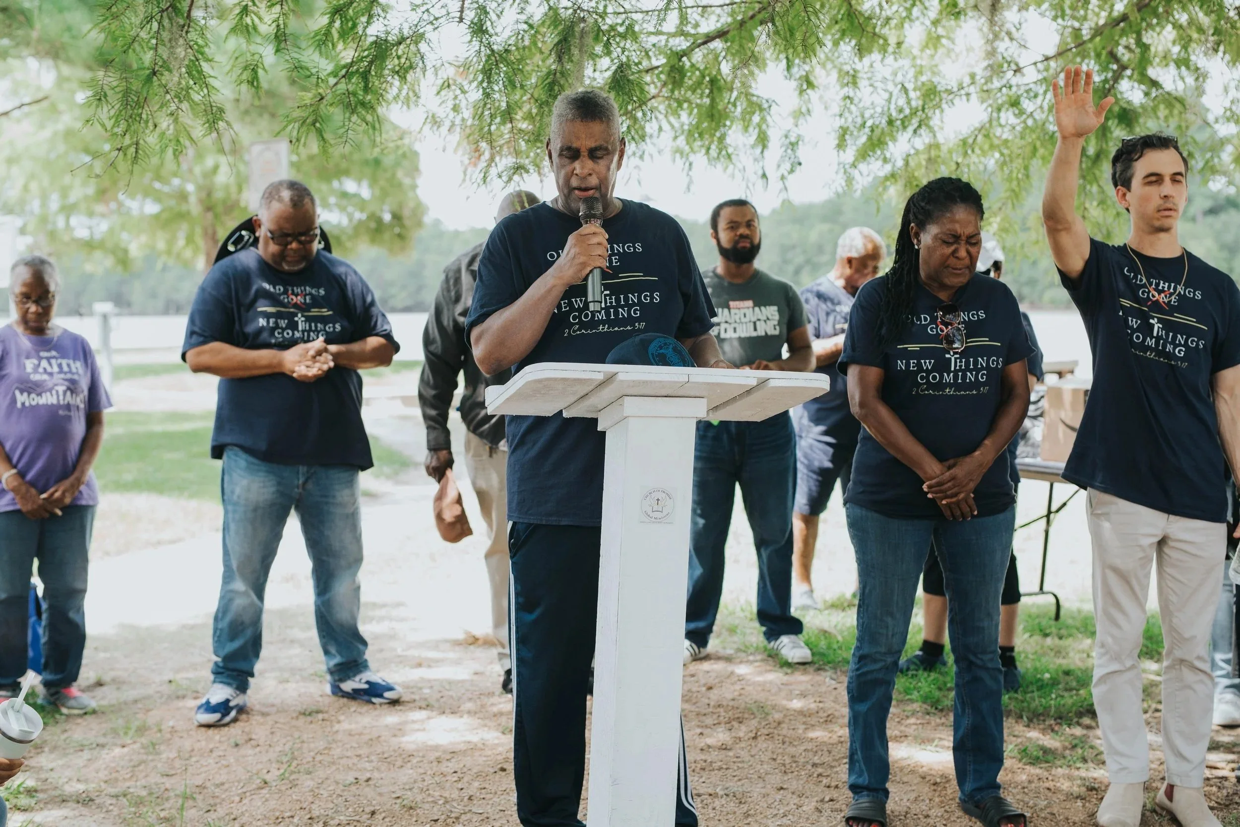 A group of people standing outdoors under a tree with green leaves, participating in a solemn prayer or moment of silence, with some people closing their eyes and one person raising a hand.