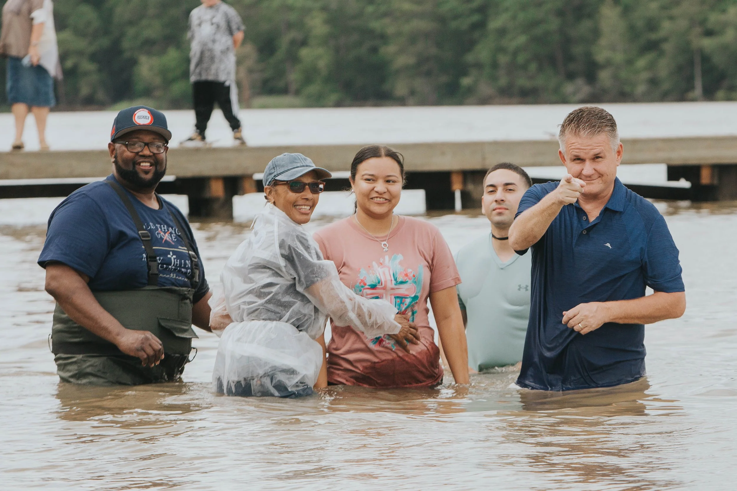 Group of five people standing in waist-deep water at a lake or river, surrounded by trees, with some on a dock in the background. They are smiling and looking toward the camera, wearing casual clothing, with one person pointing at the camera.