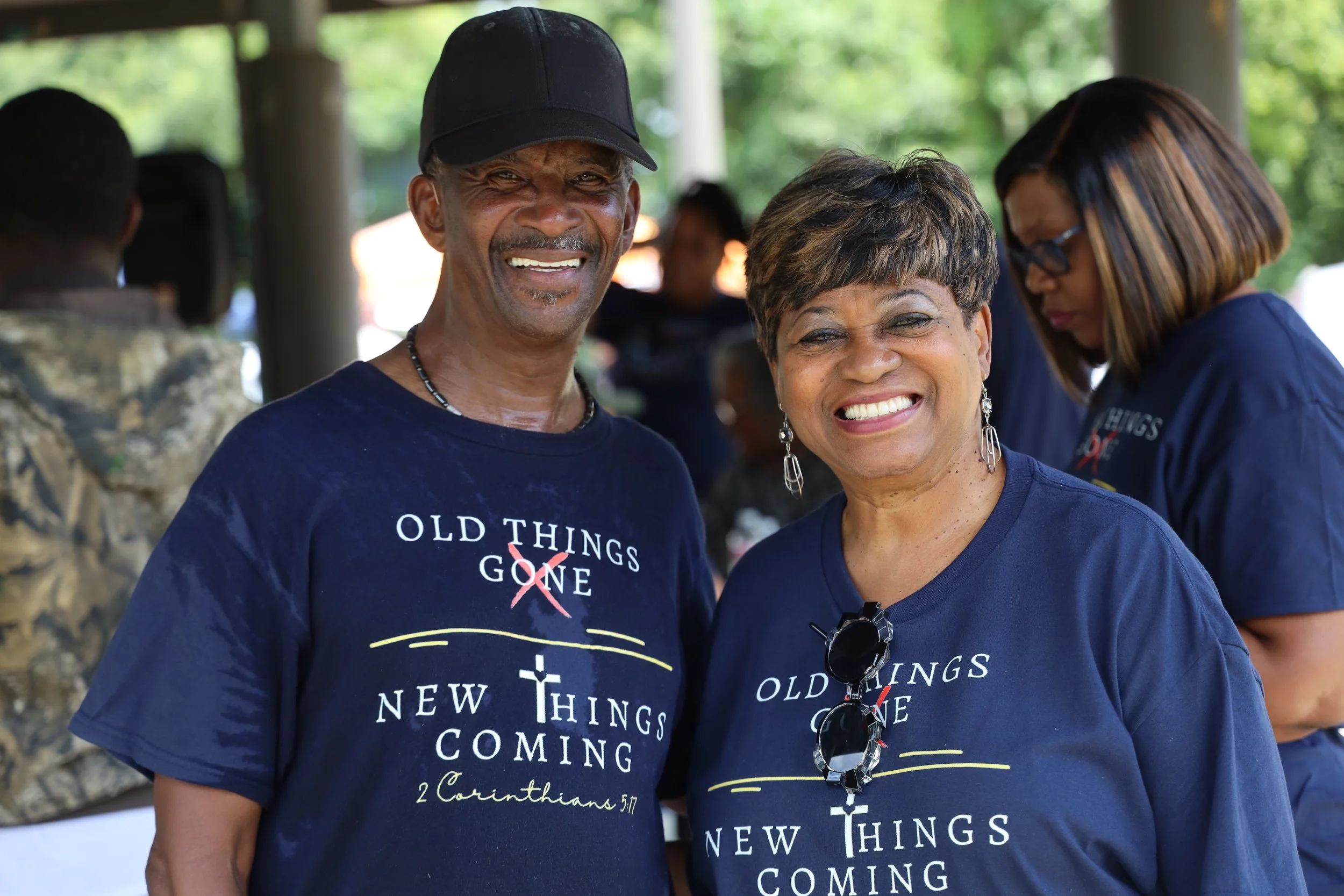 Three smiling African American people at an outdoor event wearing matching dark blue t-shirts with the words 'Old Things Gone, New Things Coming' and a cross graphic. The man on the left is wearing a black cap, and the woman in the middle has short hair and earrings. The woman on the right has glasses and straight shoulder-length hair.