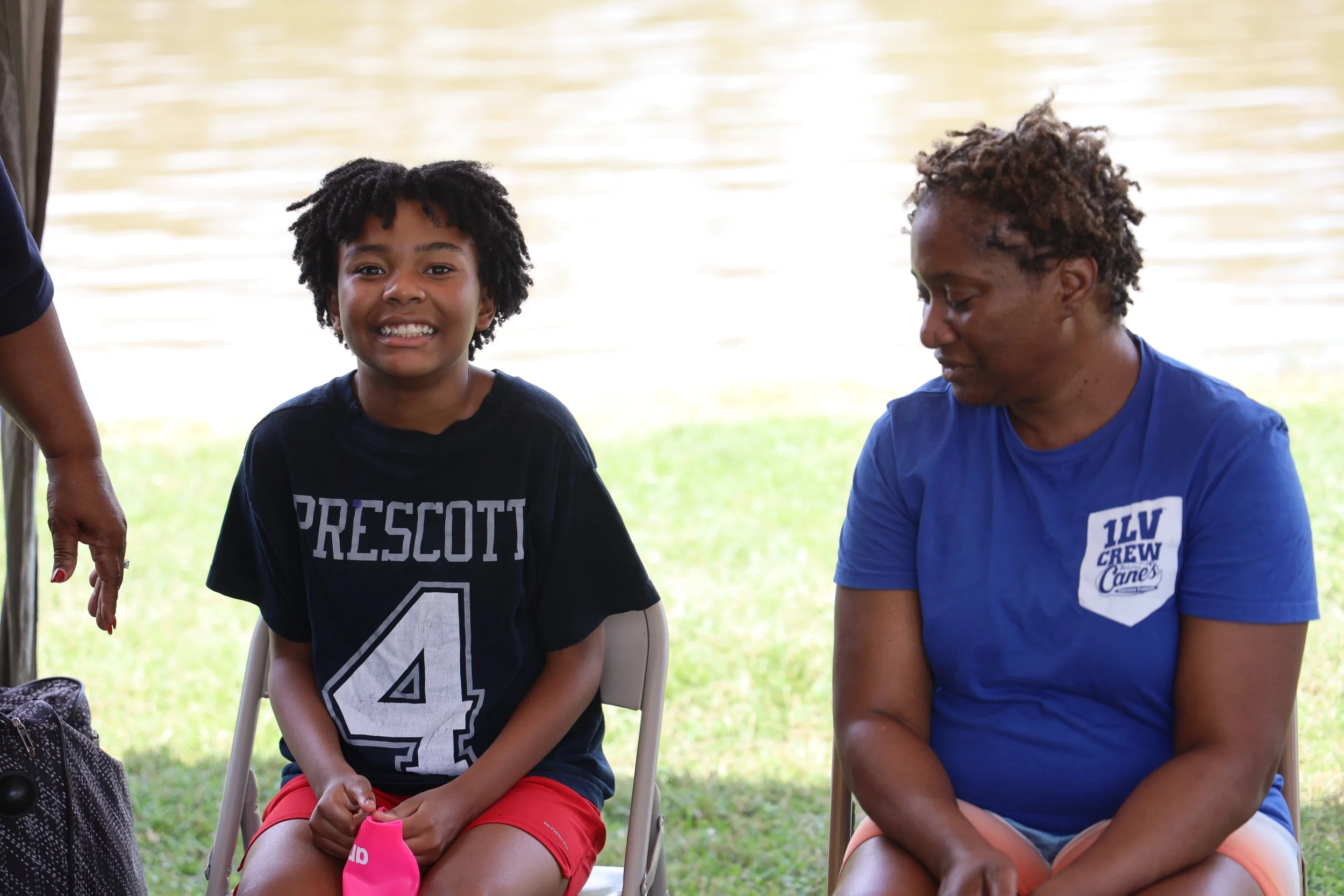 A young boy with short curly hair smiling, sitting on a chair outdoors next to a woman with short hair looking down. They are under a tent or canopy near a body of water, with grass visible in the background.