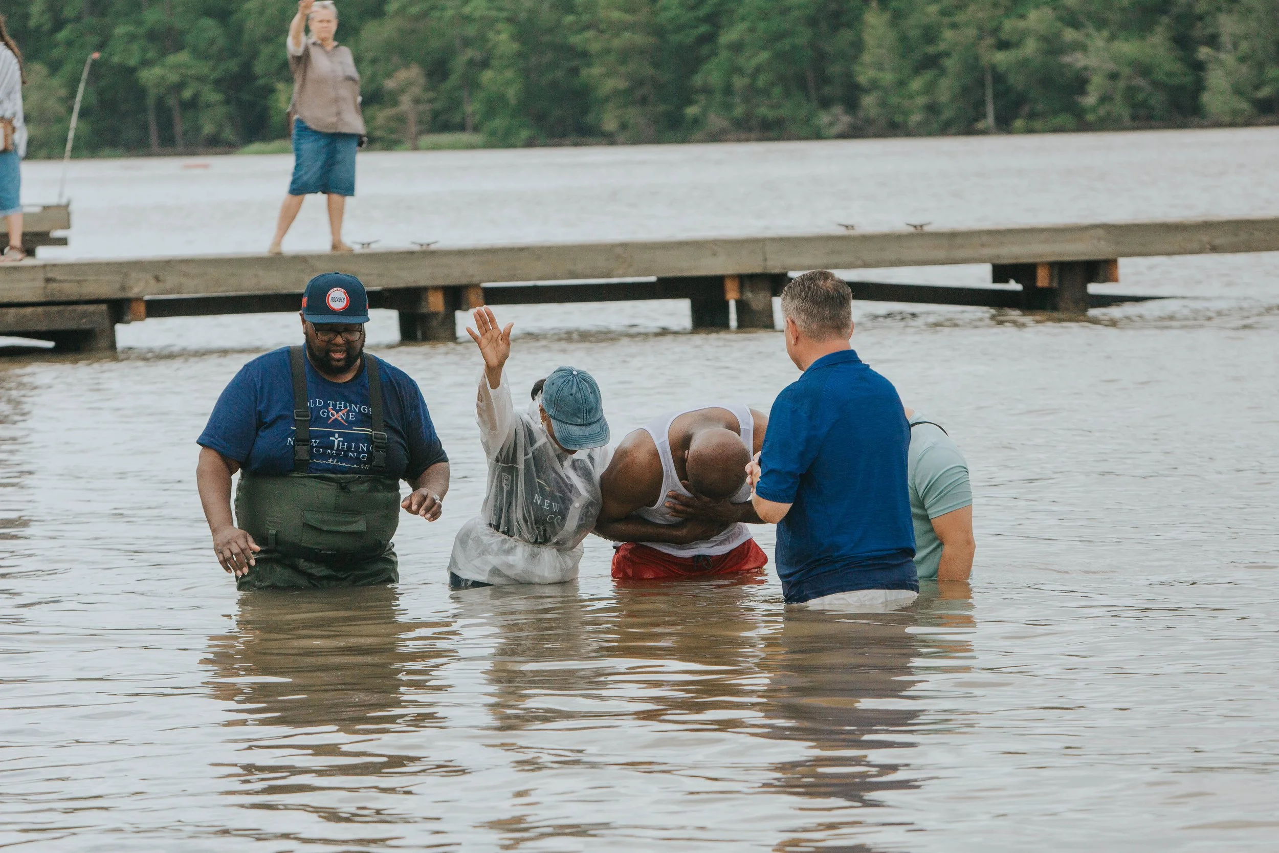 People are participating in a baptism ceremony in a river, with a group standing in the water and a dock in the background.