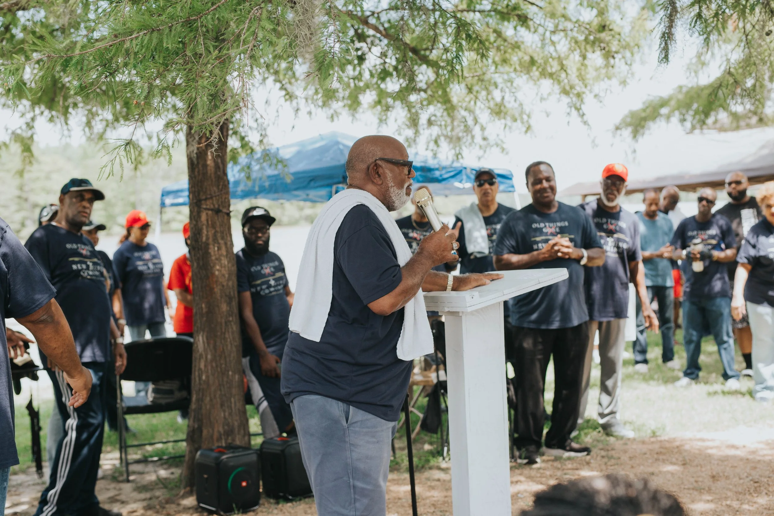 An older man with glasses and a white towel around his neck is speaking into a microphone at an outdoor gathering under a tree. Several people are standing around him, some wearing matching dark T-shirts, listening attentively.