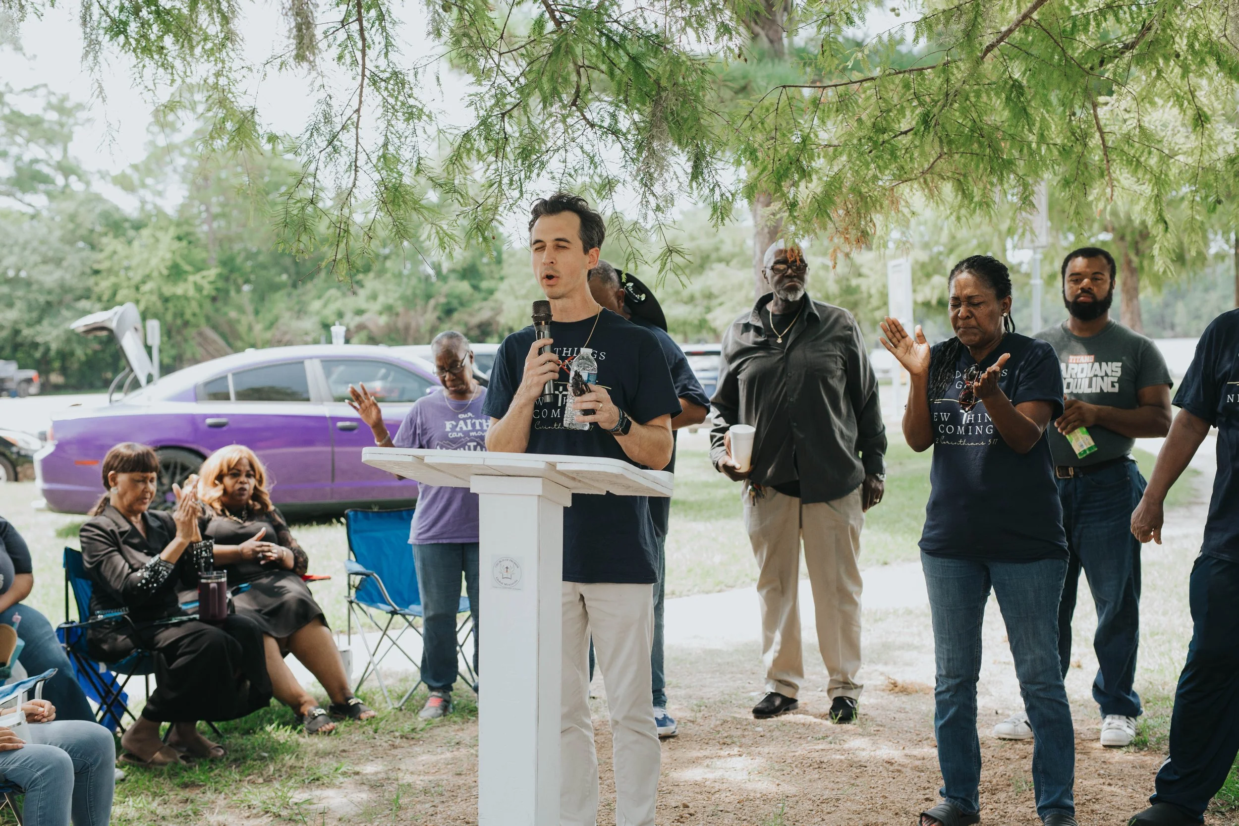 A man standing at a white podium outdoors speaking into a microphone while a group of people stand and sit around him, with some raising their hands, under green tree branches.