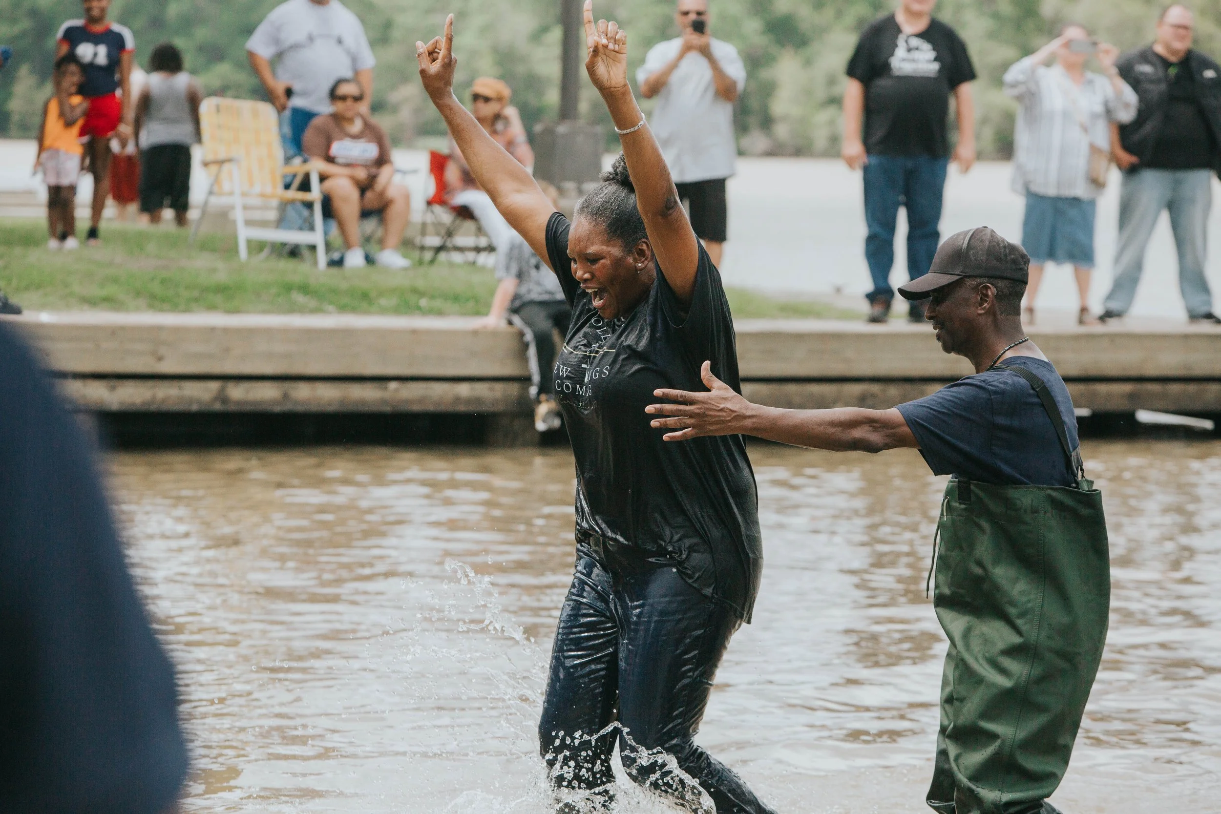 Woman being baptized in water while another person assists her in a park with people sitting and standing nearby.
