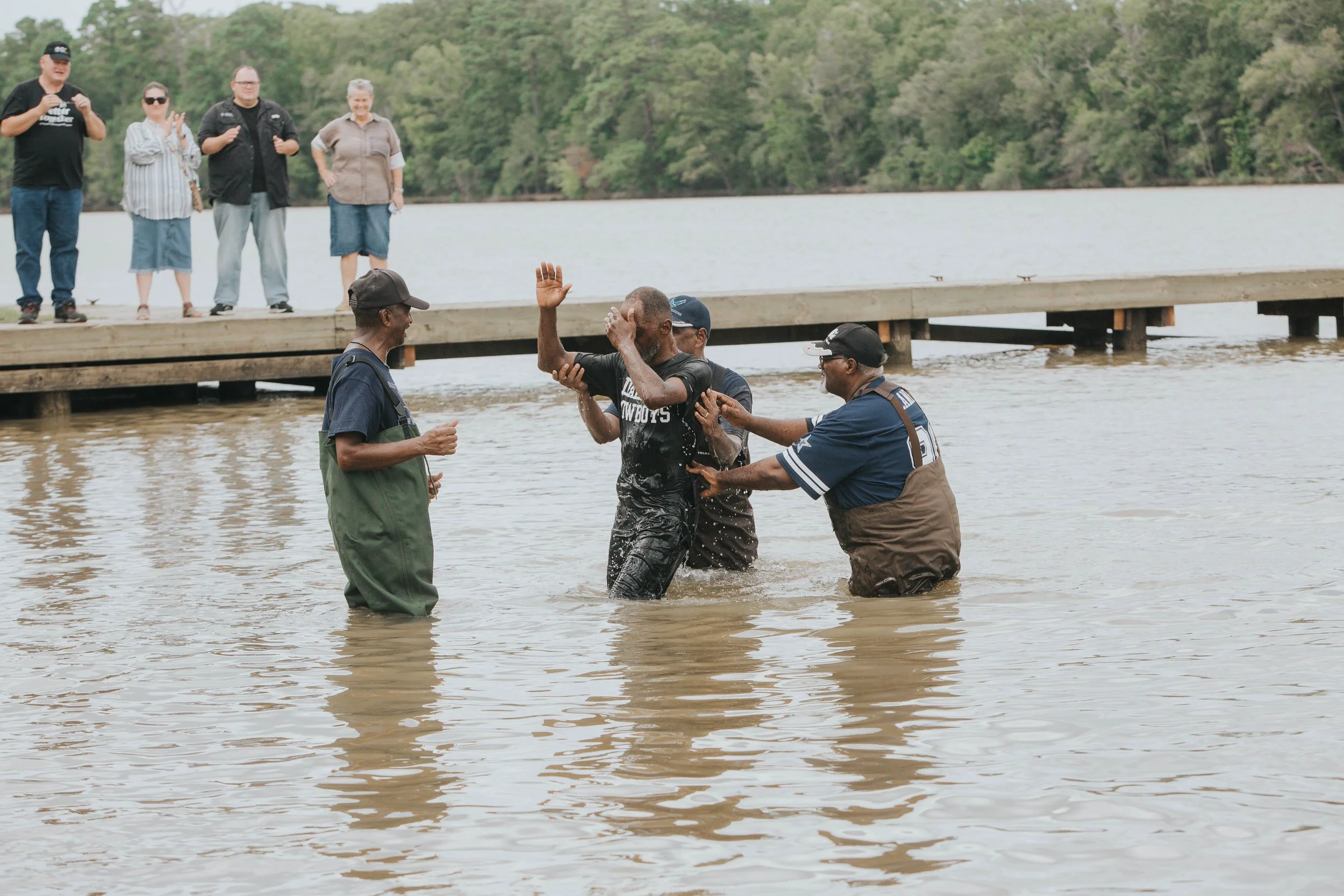 A group of people assist a man who is being baptized in a body of water near a wooden dock with trees in the background.