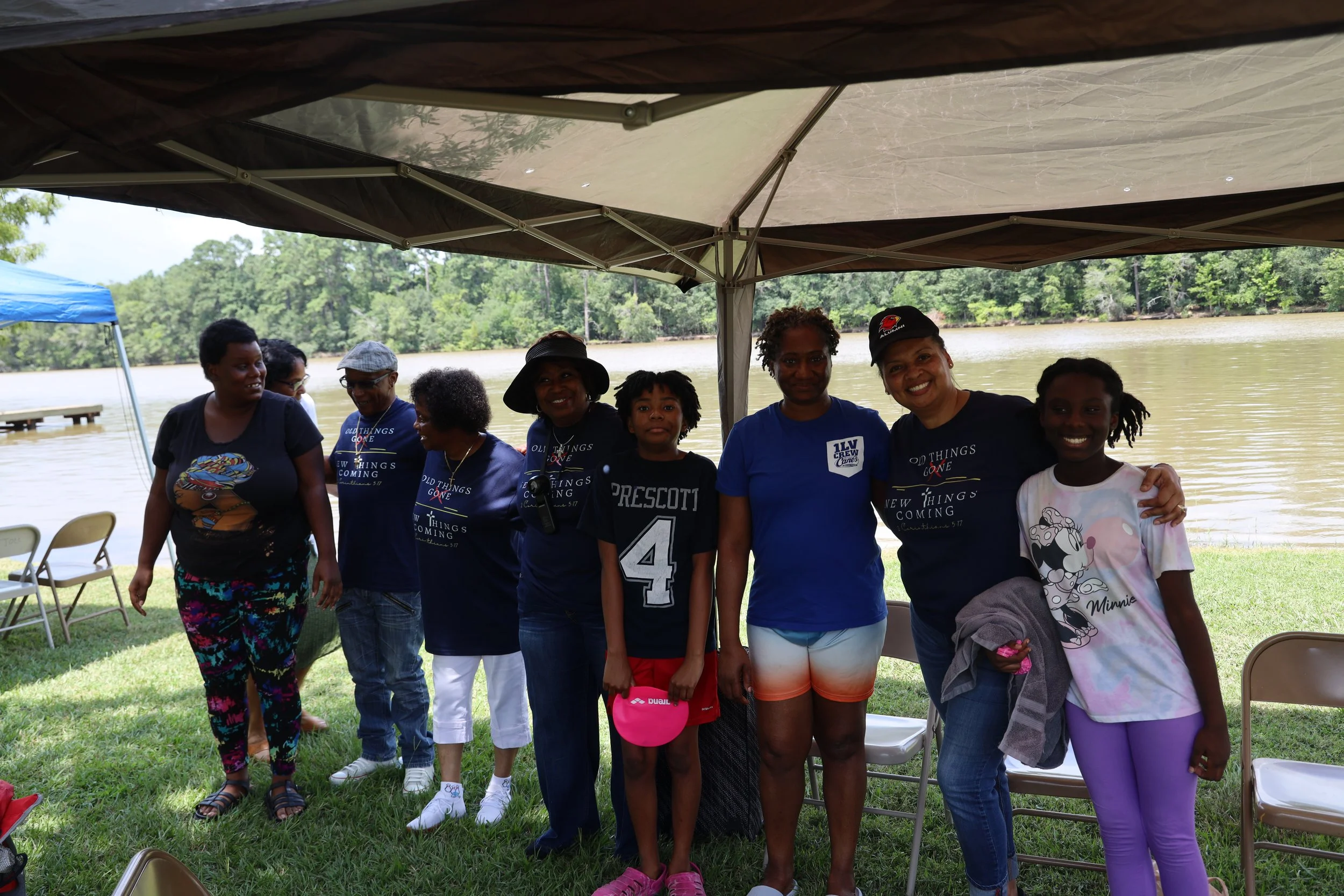 Group of nine women and girls standing under a canopy by a lake, smiling at the camera, with chairs nearby and trees in the background.