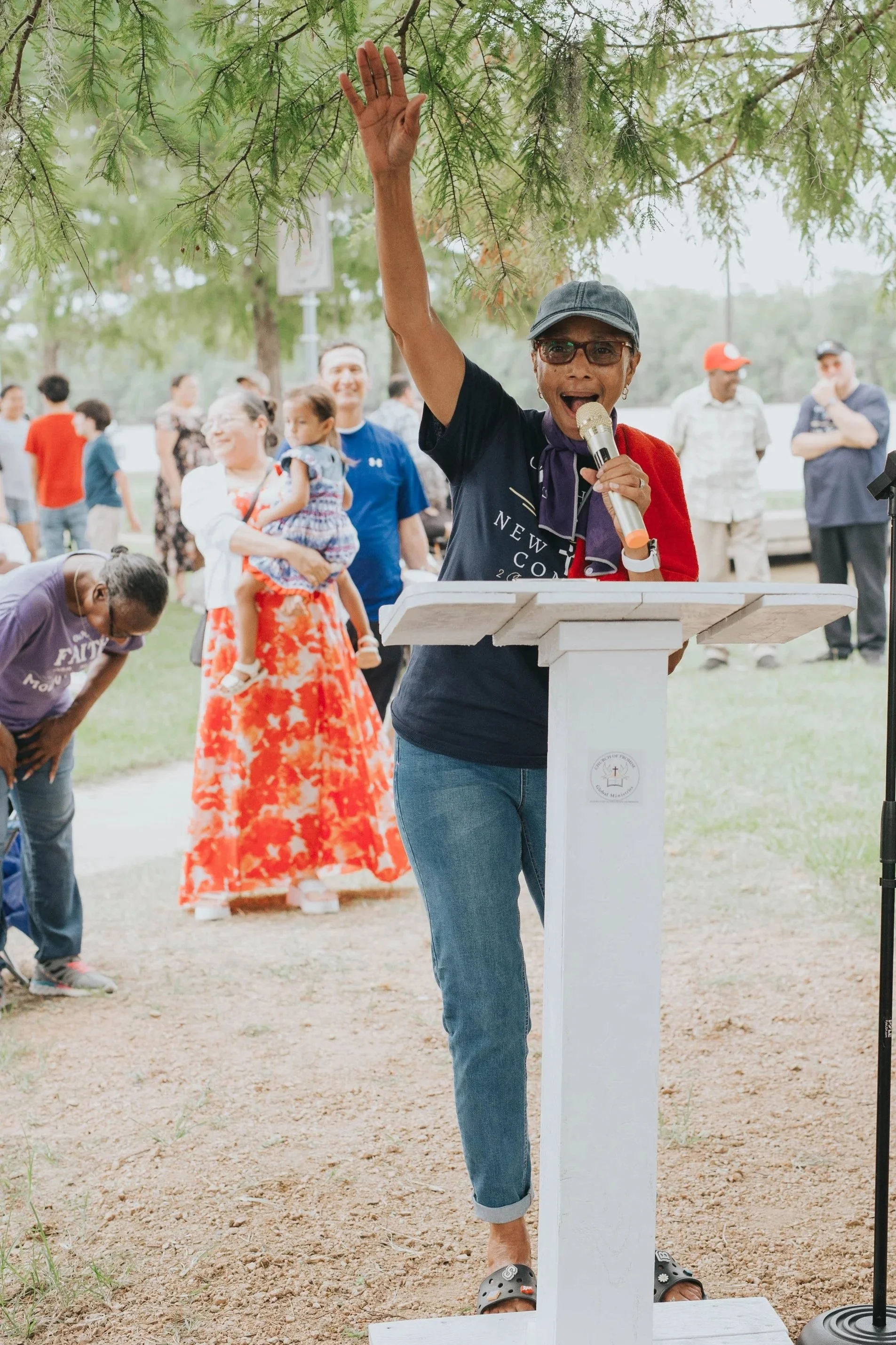 Woman speaking at outdoor event, raising her hand, standing at a white podium, with a crowd of diverse people in the background under trees.